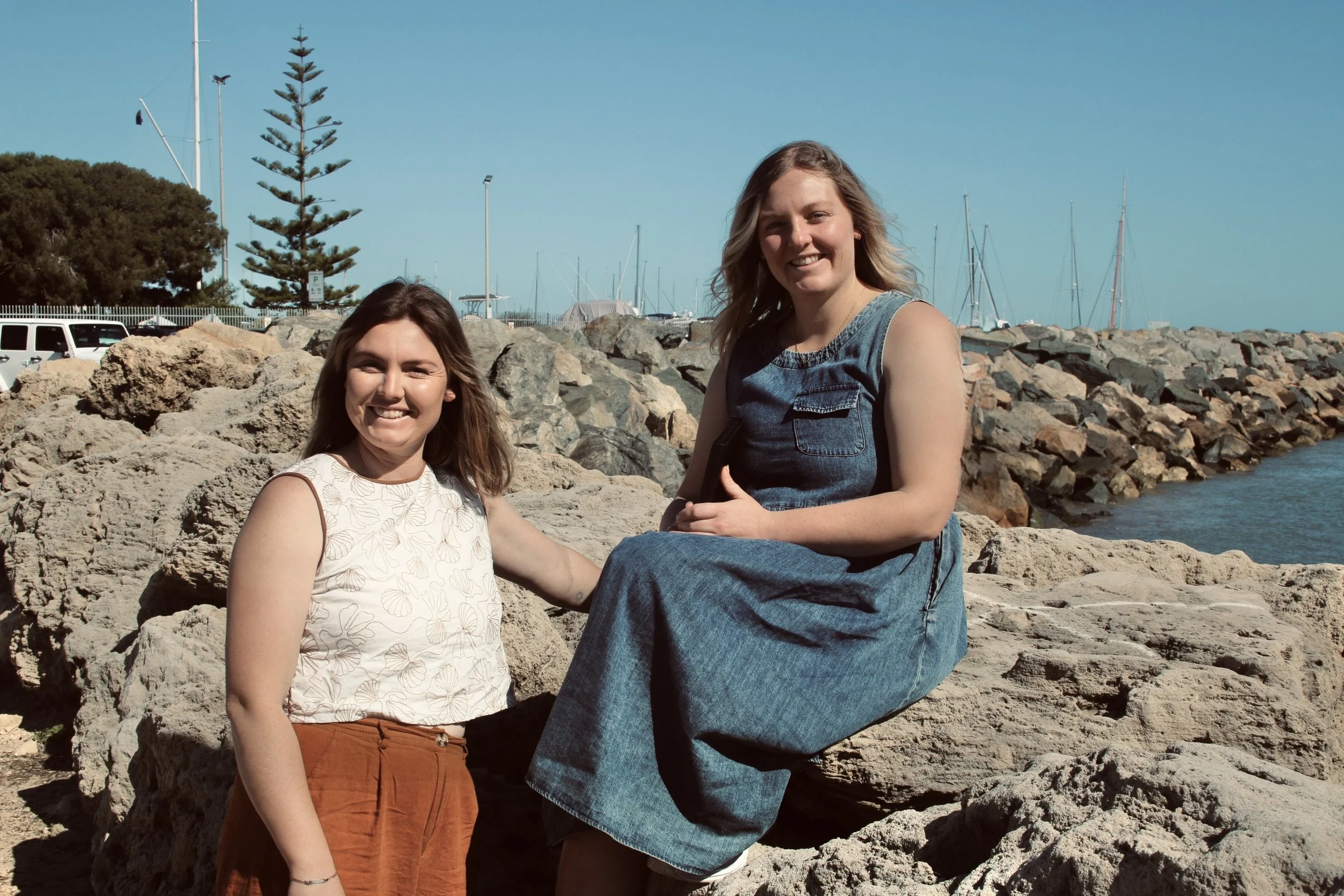 Two young women smiling on rocks near a marina with sailboats, clear blue sky, and a pine tree in the background.
