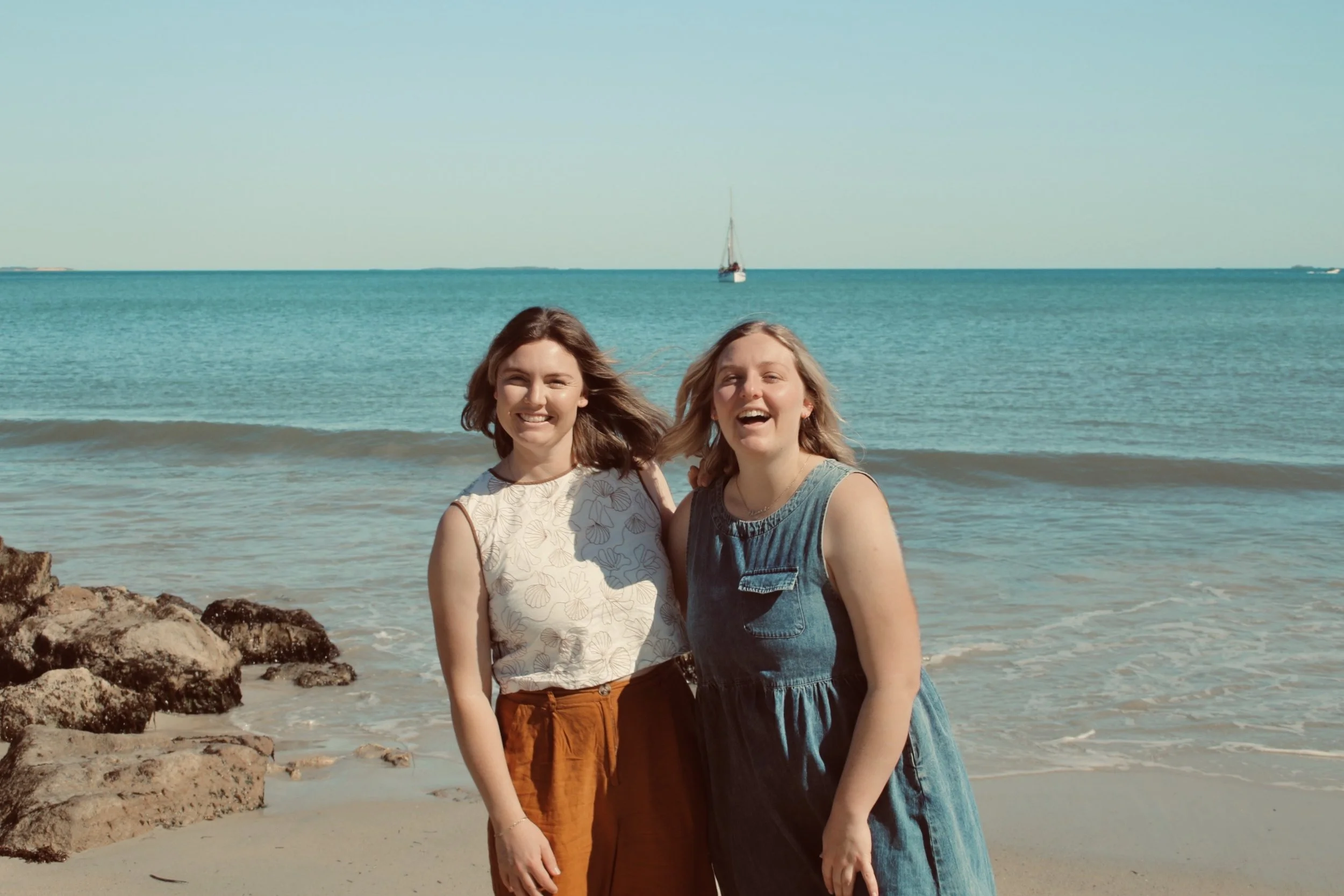 Two smiling women stand on the beach near rocks, with the ocean and a sailboat in the background on a sunny day.