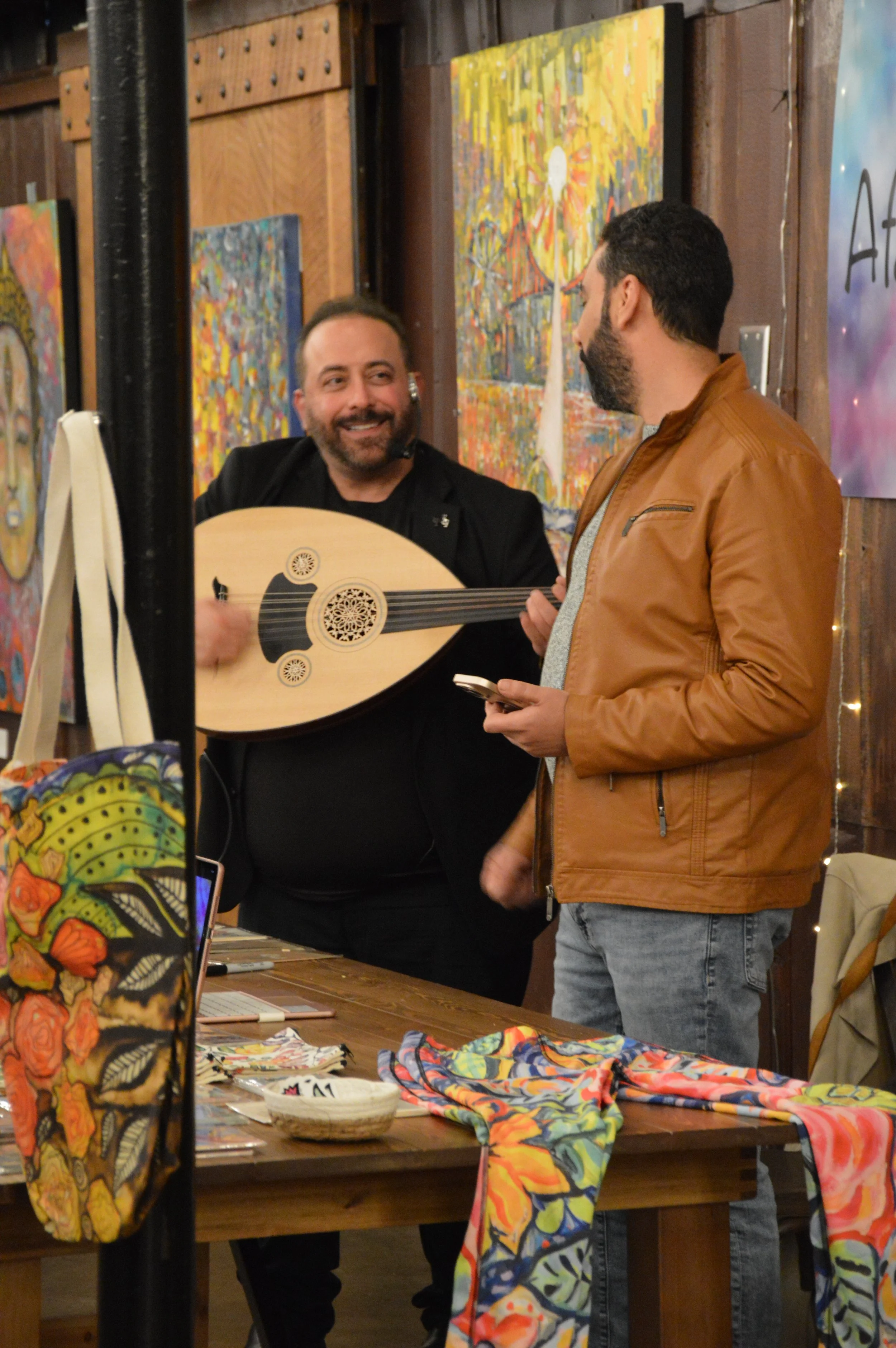 Iraqi musician and oud player Laith Alattar at Middle Eastern Arts Collective Inaugural Art Gallery next to paintings by Egyptian visual artist Afaf Khalil