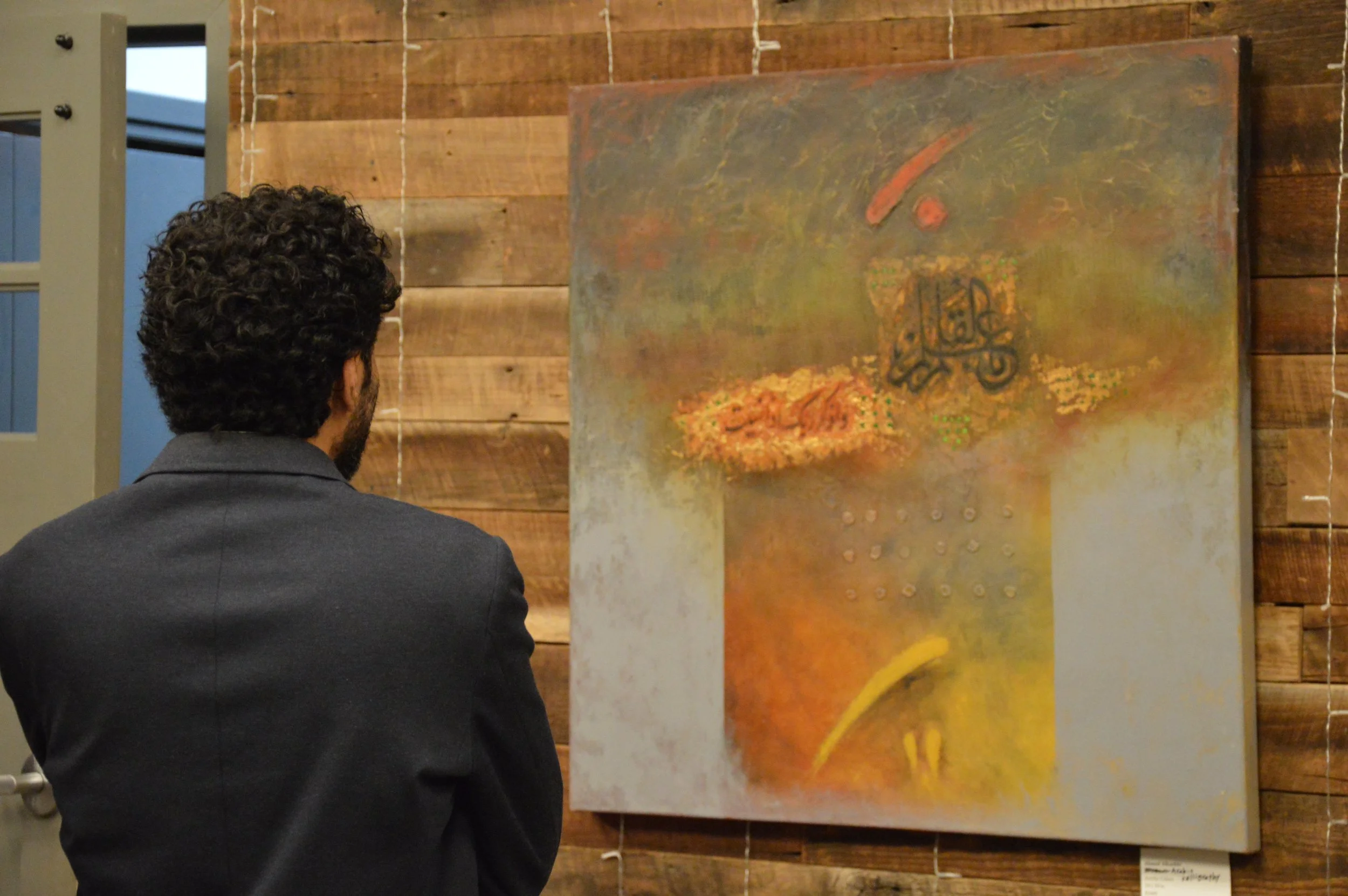 Attendee of the Middle Eastern Arts Collective's inaugural art gallery examining a painting by Iraqi artist Ahmed Alkarkhi. Painting is grey, yellow, orange, and green with arabic letters and abstract composition.
