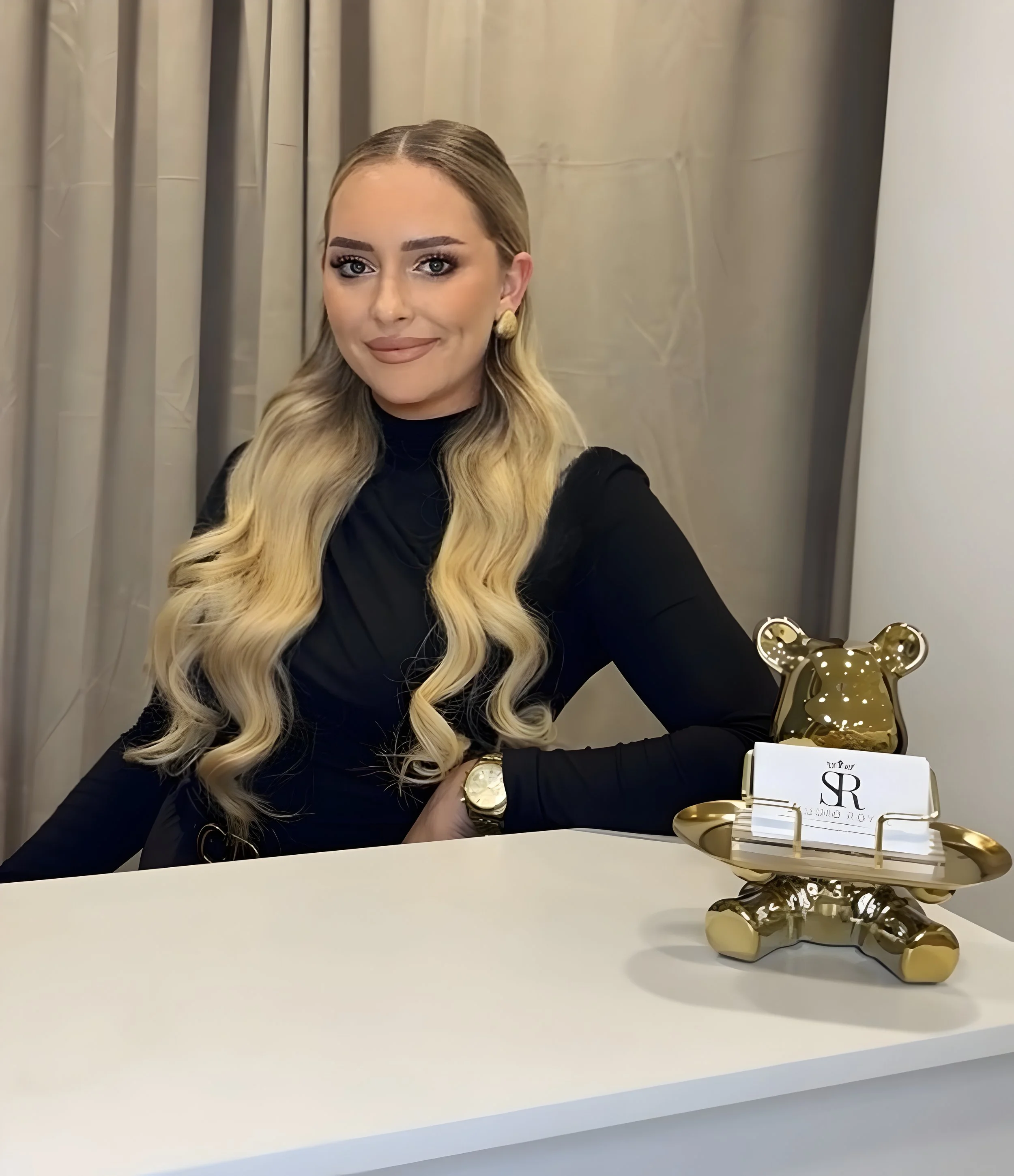Eine elegante Frau mit langen, gewellten blonden Haaren sitzt an einem weißen Tisch mit einer goldenen Teddy-Figur und einem Kartenhalter, auf dem ein Visitenkartenset steht.