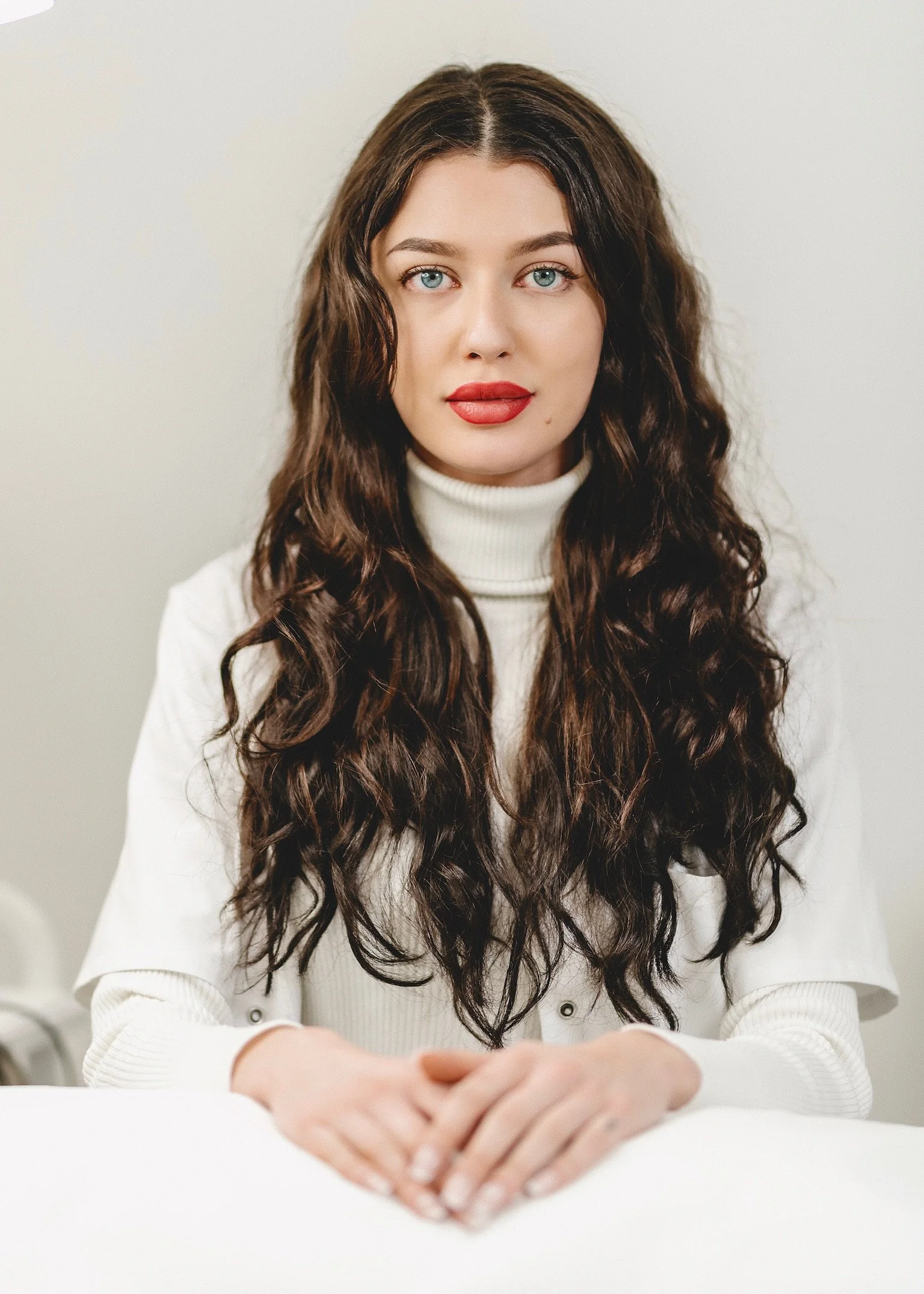 Junge Frau mit langen, lockigen braunen Haaren, rotem Lippenstift und blauen Augen, trägt einen weißen Rollkragenpullover, sitzt an einem weißen Tisch, Blick geradeaus.
