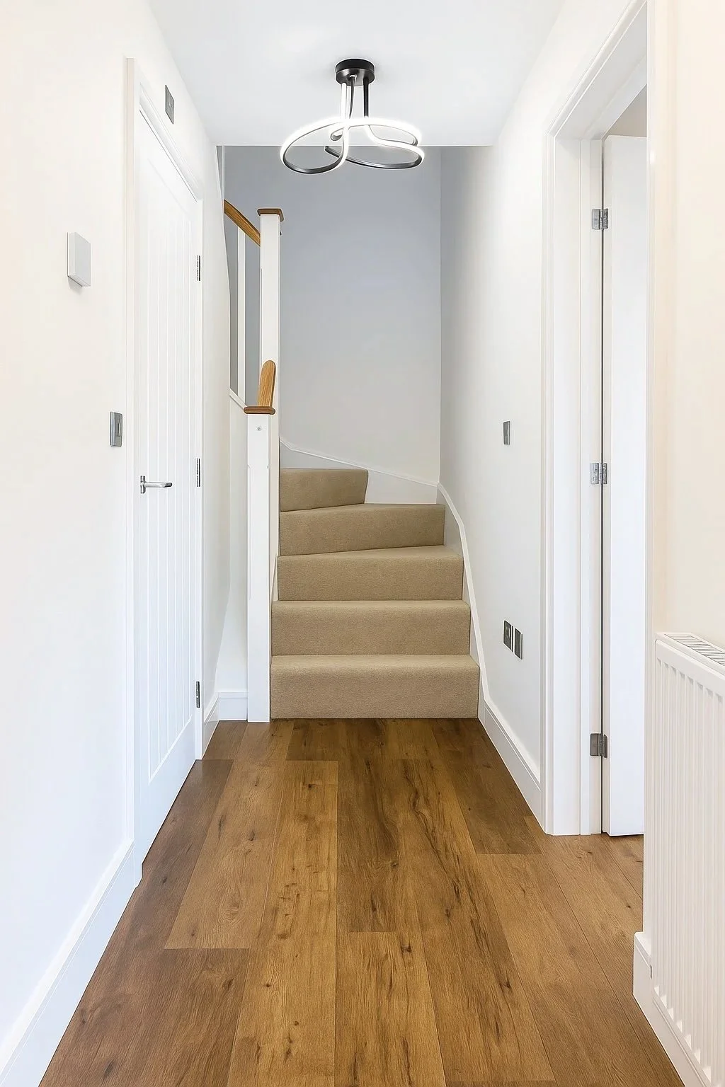 Interior view of a modern house entryway with a staircase, wooden floor, white walls, a contemporary ceiling light fixture, and doors on each side.
