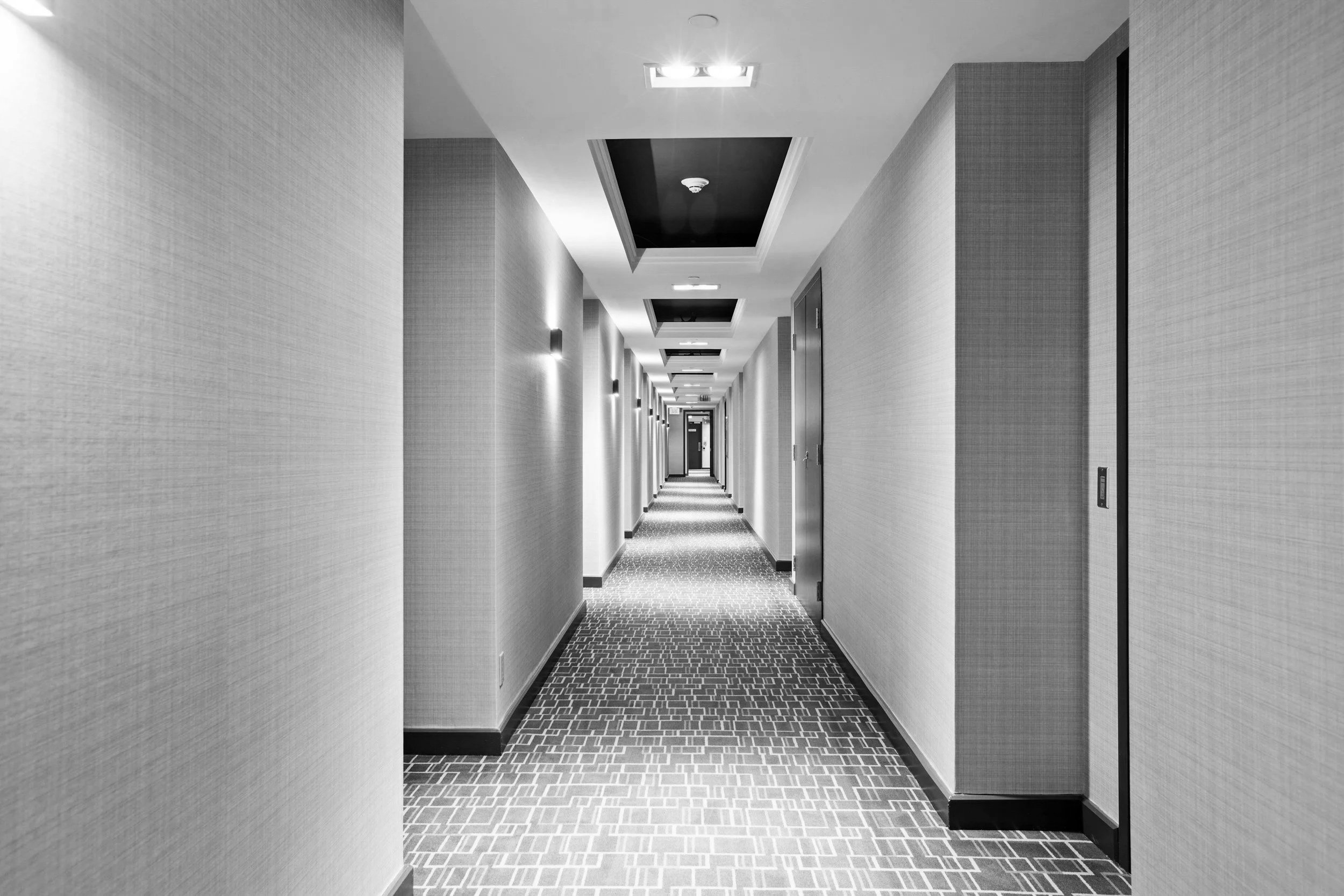 Empty hotel corridor with patterned carpet, plain walls, and ceiling lights.