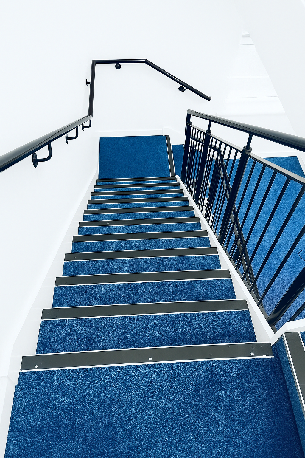 Indoor staircase with black handrails, blue carpeted steps, white walls, and a corner landing.