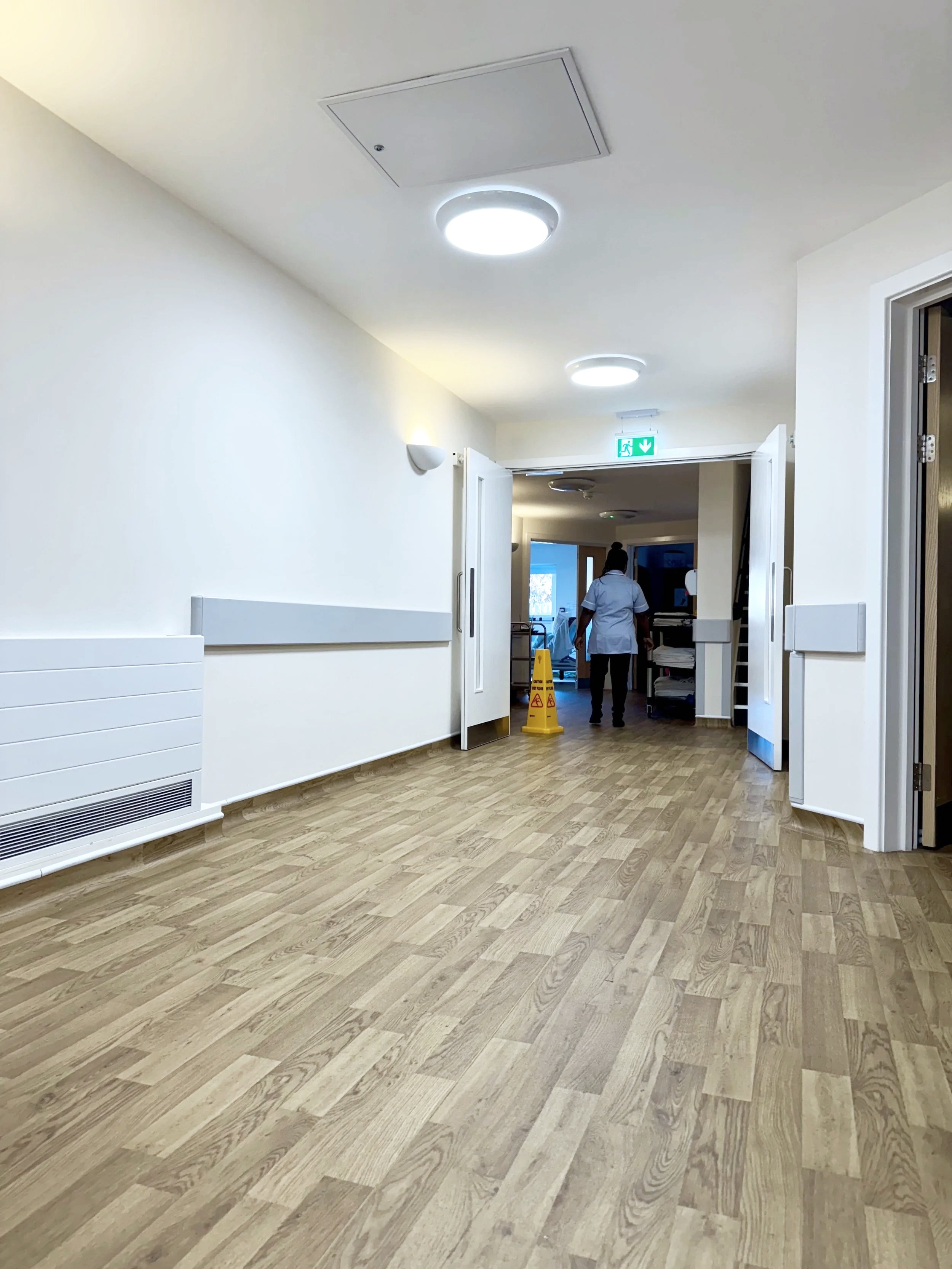 An empty hospital room with wood flooring, white walls, a wall-mounted heater, ceiling lights, a partially open door, and a person walking away in the background near a yellow caution sign.