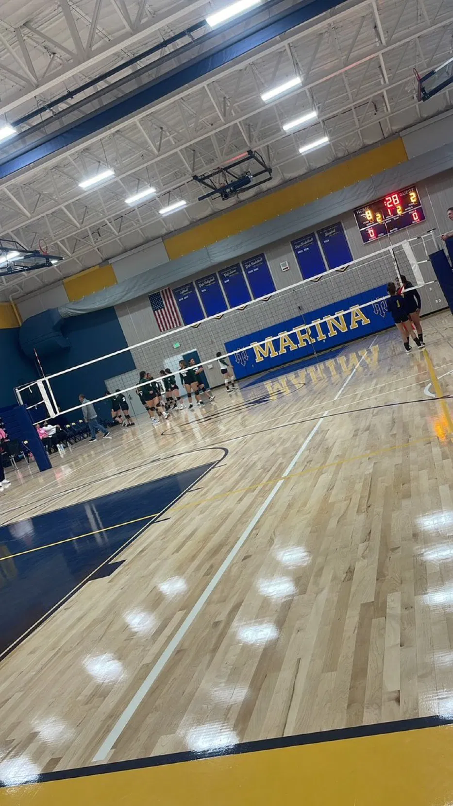 Indoor volleyball court at Marina, with players preparing for a match and a scoreboard showing 28-2 scores.