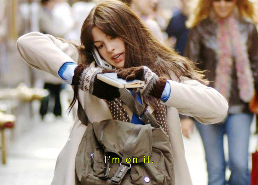 A young woman with long brown hair, wearing a coat and gloves, is talking on a cell phone while holding a notebook and a pen. She has a backpack on her back and appears to be in a busy outdoor setting with other people in the background.