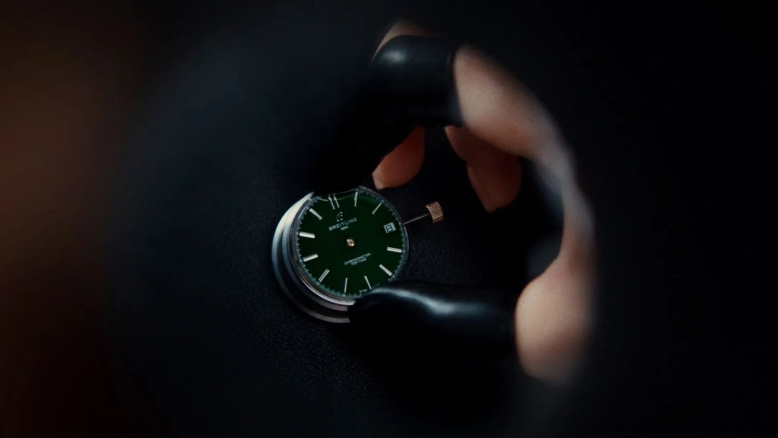 Close-up of a hand wearing a black glove holding a Breitling chronometer watch, with a green watch face and a date display, against a dark background.