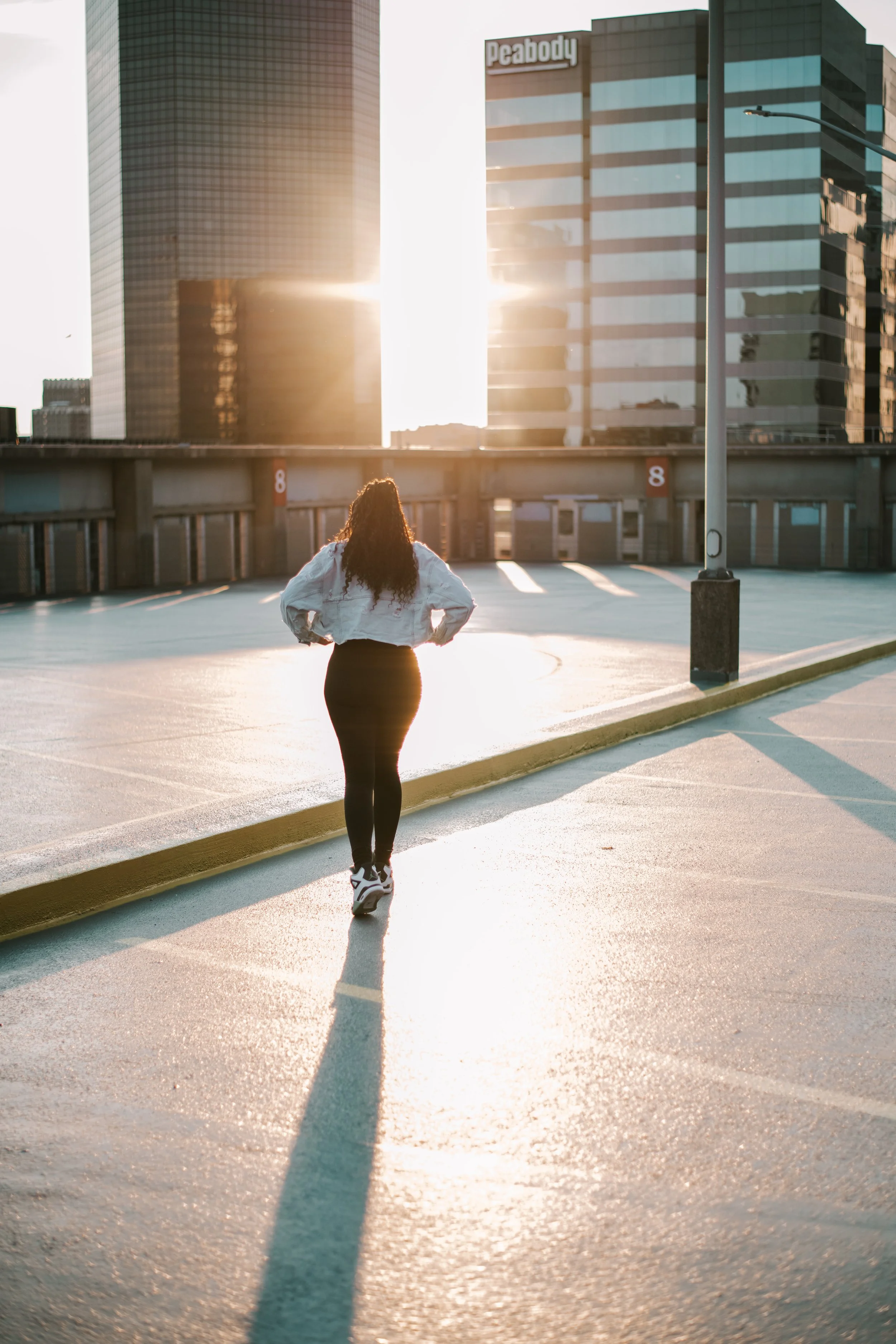 Woman with long curly hair walking in an empty parking lot during sunset, with tall modern buildings in the background.