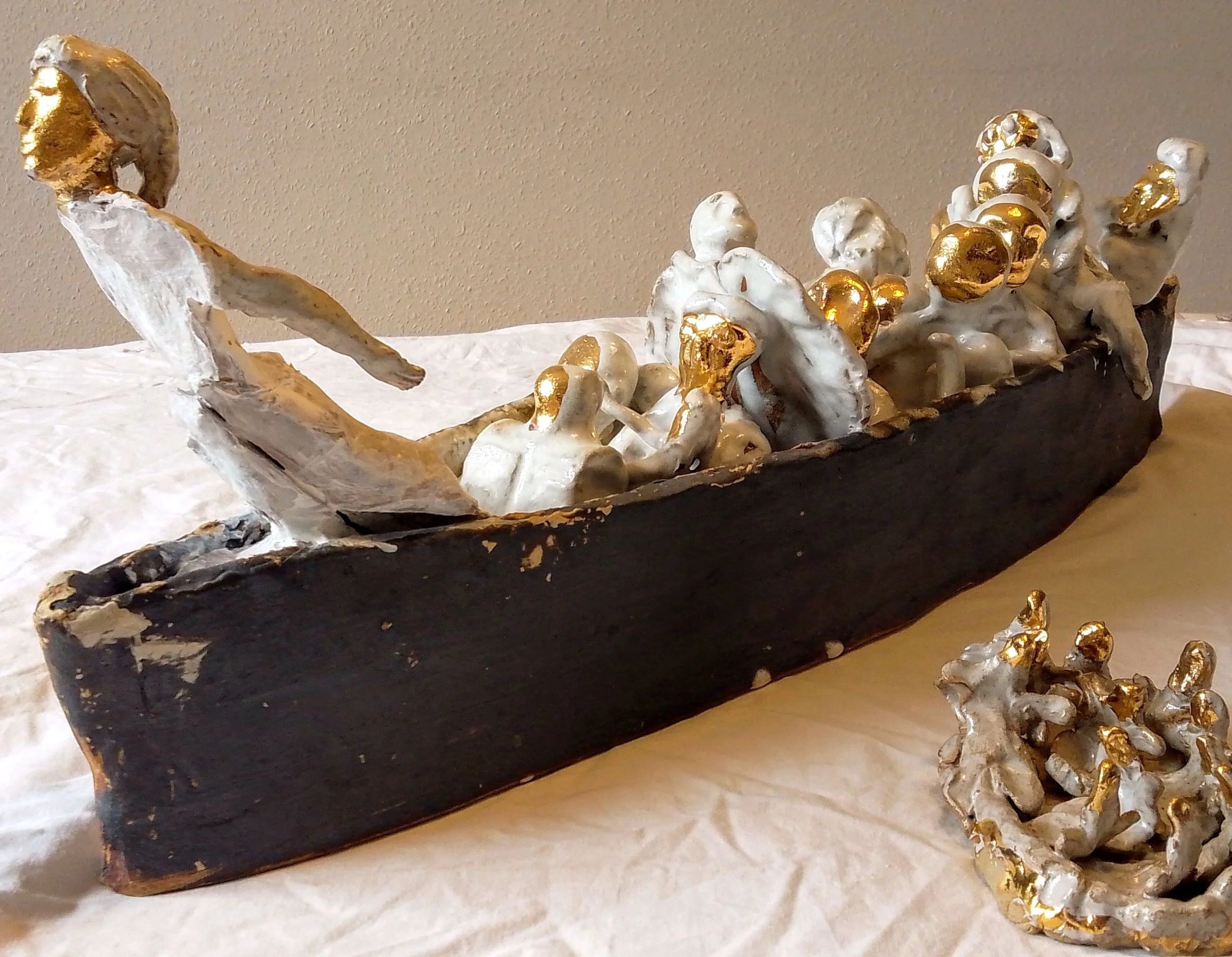 An abstract sculpture of a boat with human figures, painted in white and gold, with a black base, displayed on a white cloth.