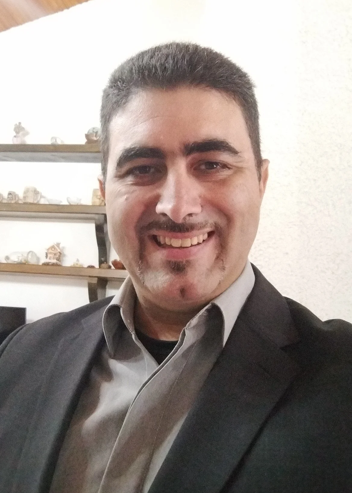 A man smiling, wearing a black suit and light gray shirt, indoors with shelves of decorative items in the background.