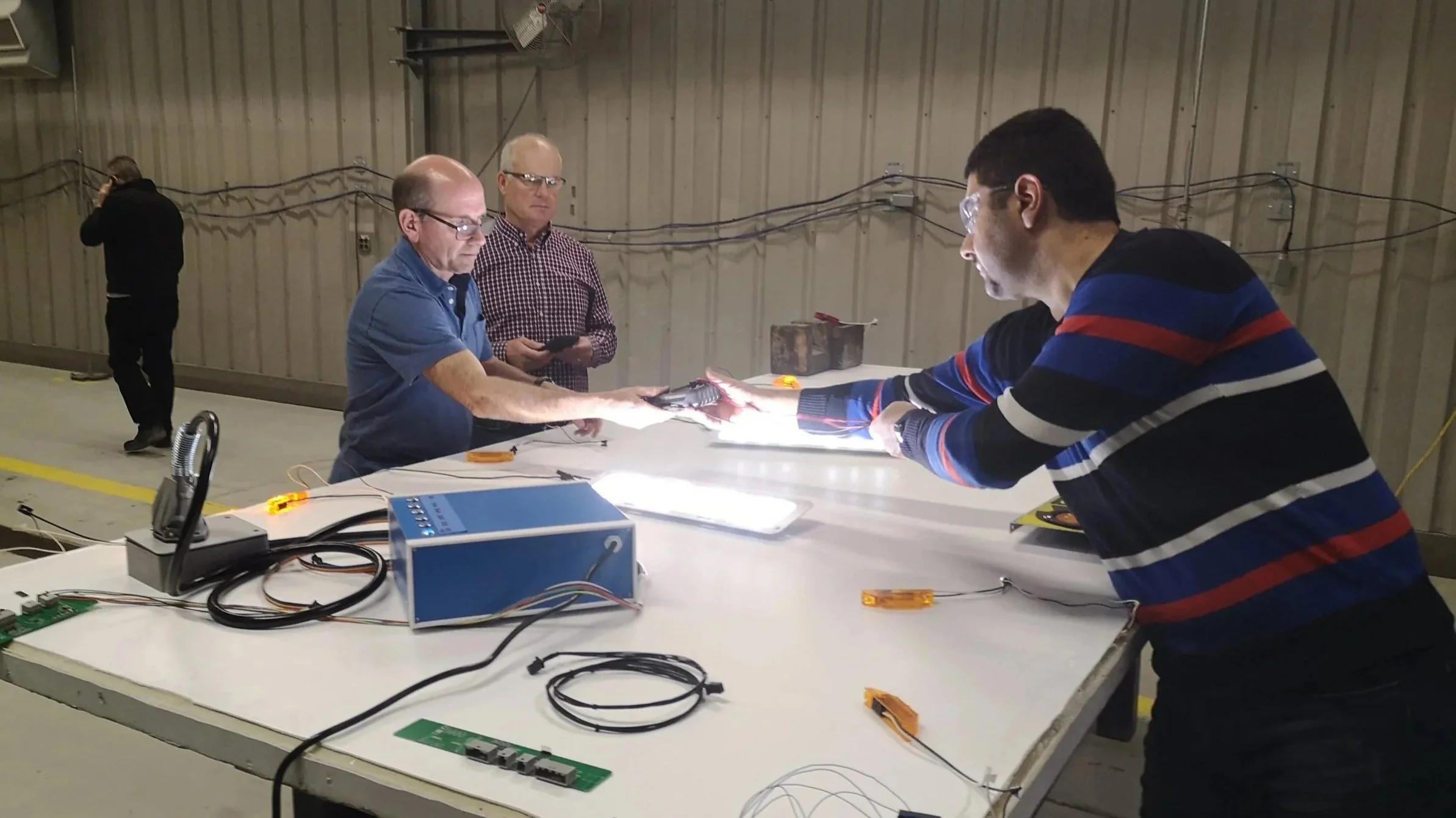 Two men are working on an electronics project at a white work table, with one man handing an object to the other while a third man observes, all in an industrial setting.