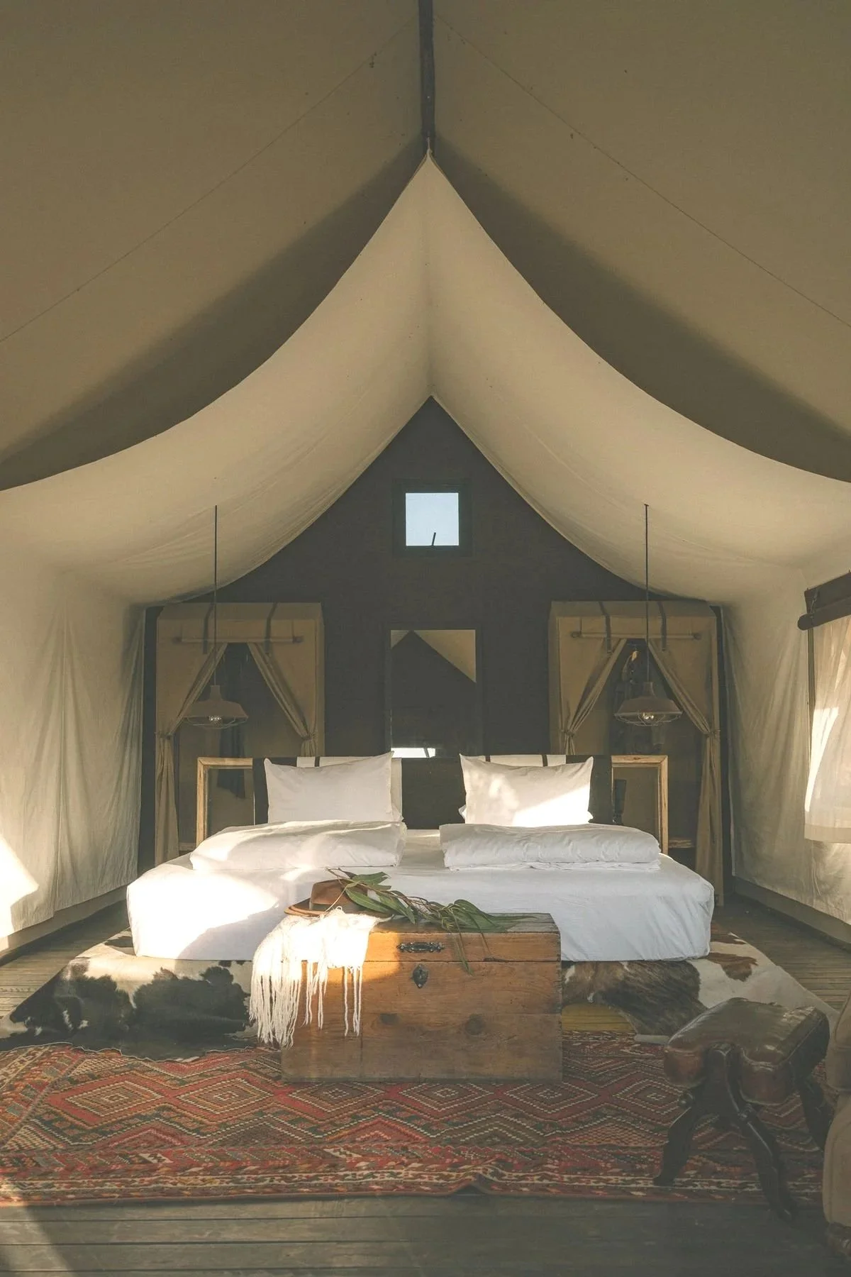 Inside a spacious, rustic tent with a large bed featuring white linens, two pillows, and a wooden chest at the foot of the bed. The tent has canvas walls and a high, peaked ceiling, with windows and curtains. The floor is wooden, with a colorful woven rug in front of the bed and a small wooden stool nearby.