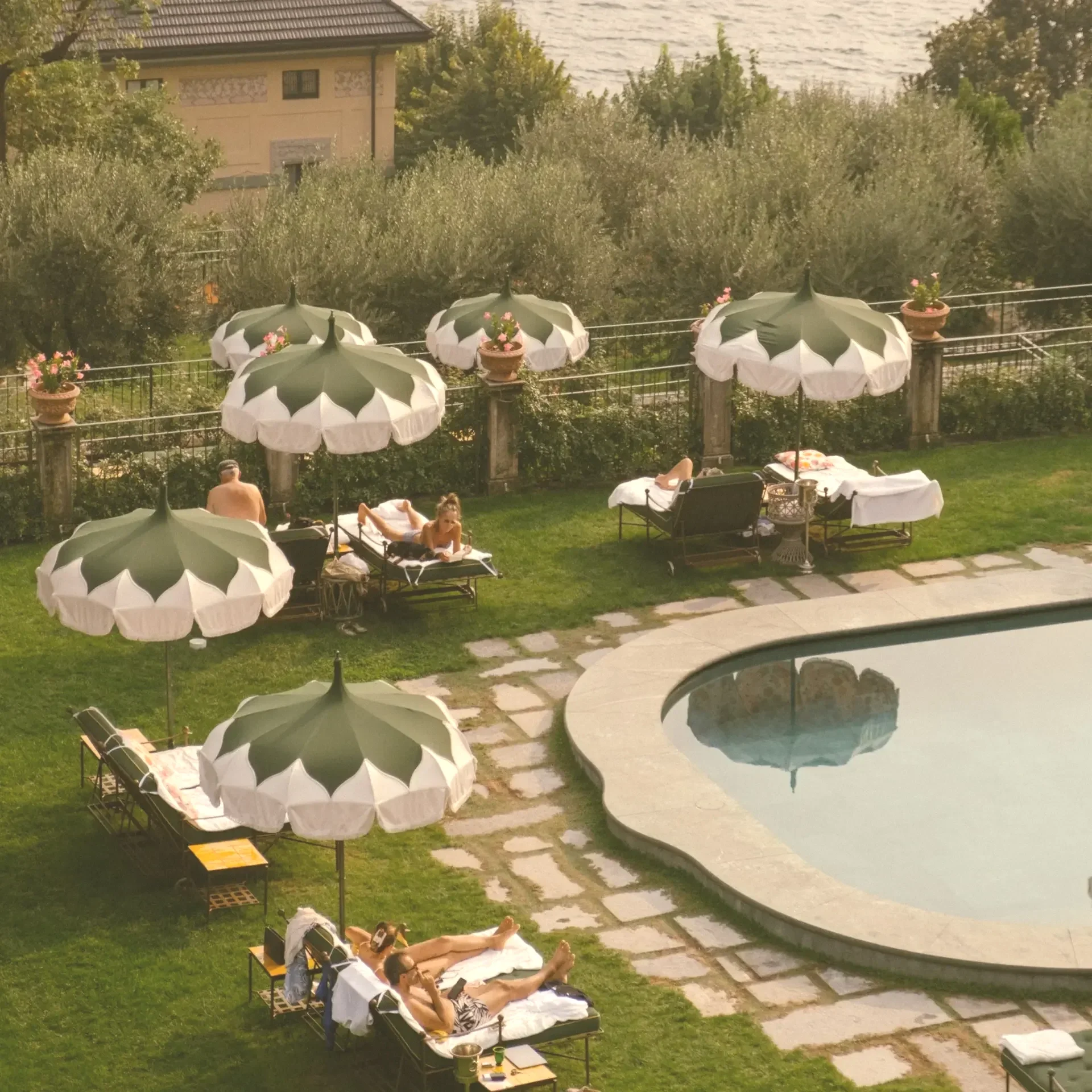 People relaxing by a swimming pool with umbrellas and lounge chairs in a lush backyard.