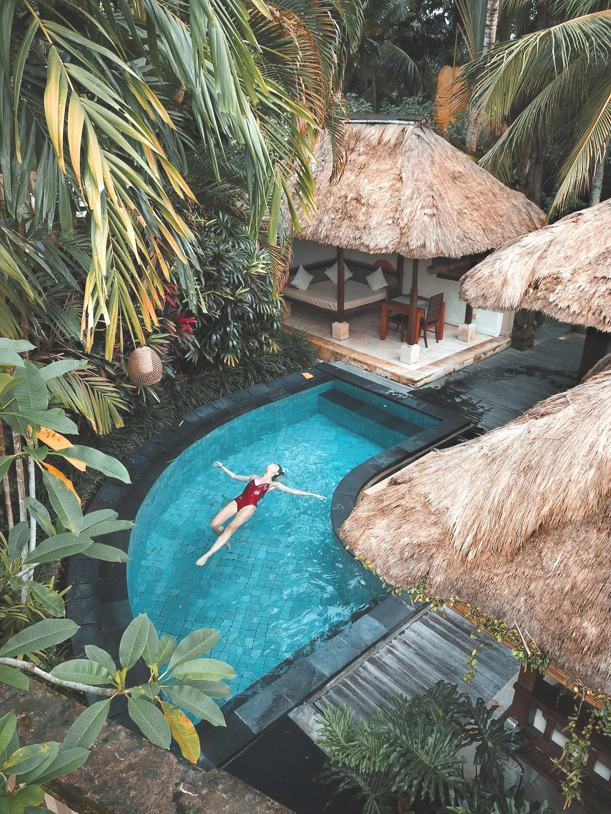 A woman in a red swimsuit floating in a small, curved swimming pool surrounded by tropical plants and thatched-roof huts.