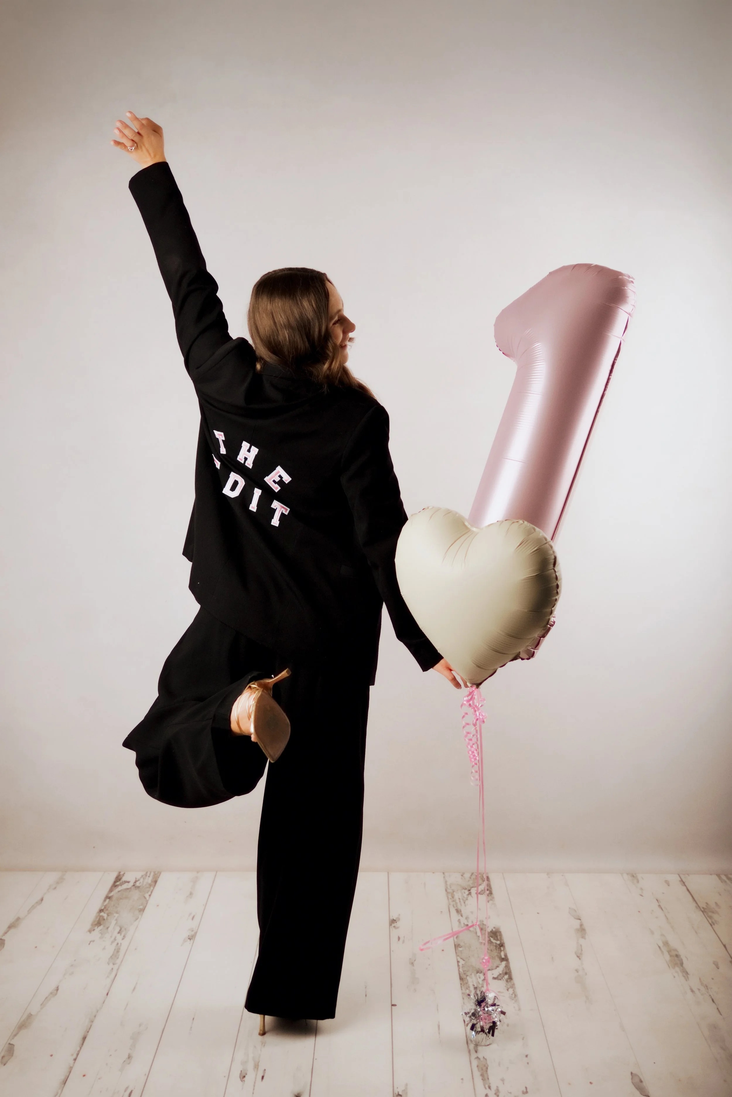 A woman dressed in black with a long black skirt, black blazer, and beige high heels, is posing with one knee bent and one arm raised. She is holding a large, metallic pink heart-shaped balloon and a long, thin pink string with silver and pink tinsel decorations at the end. The background is plain, and the floor is a white wooden surface with some weathered areas.