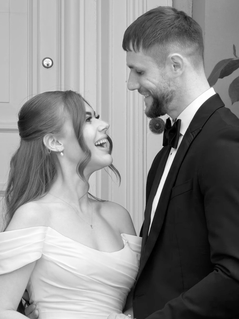 Black and white photo of a woman and man smiling and looking at each other, dressed in wedding attire, standing indoors.