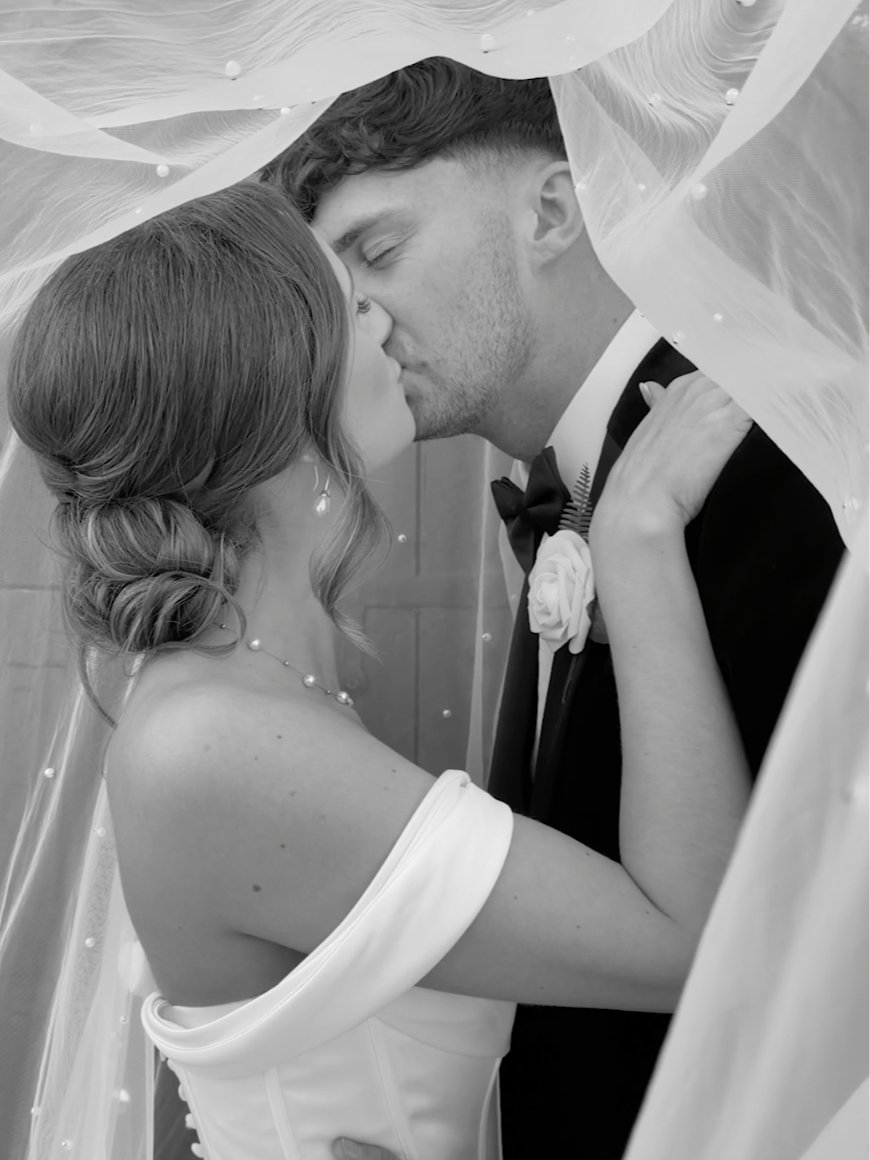 A bride and groom share a kiss under a sheer veil, with the bride wearing an off-the-shoulder wedding dress and pearl jewelry, and the groom in a tuxedo with a boutonnière.