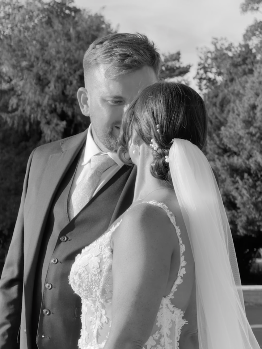 Black and white photo of a groom and bride close together during their wedding, outdoors with trees in the background. The groom is wearing a suit with a tie, and the bride is wearing a lace wedding dress with a veil.