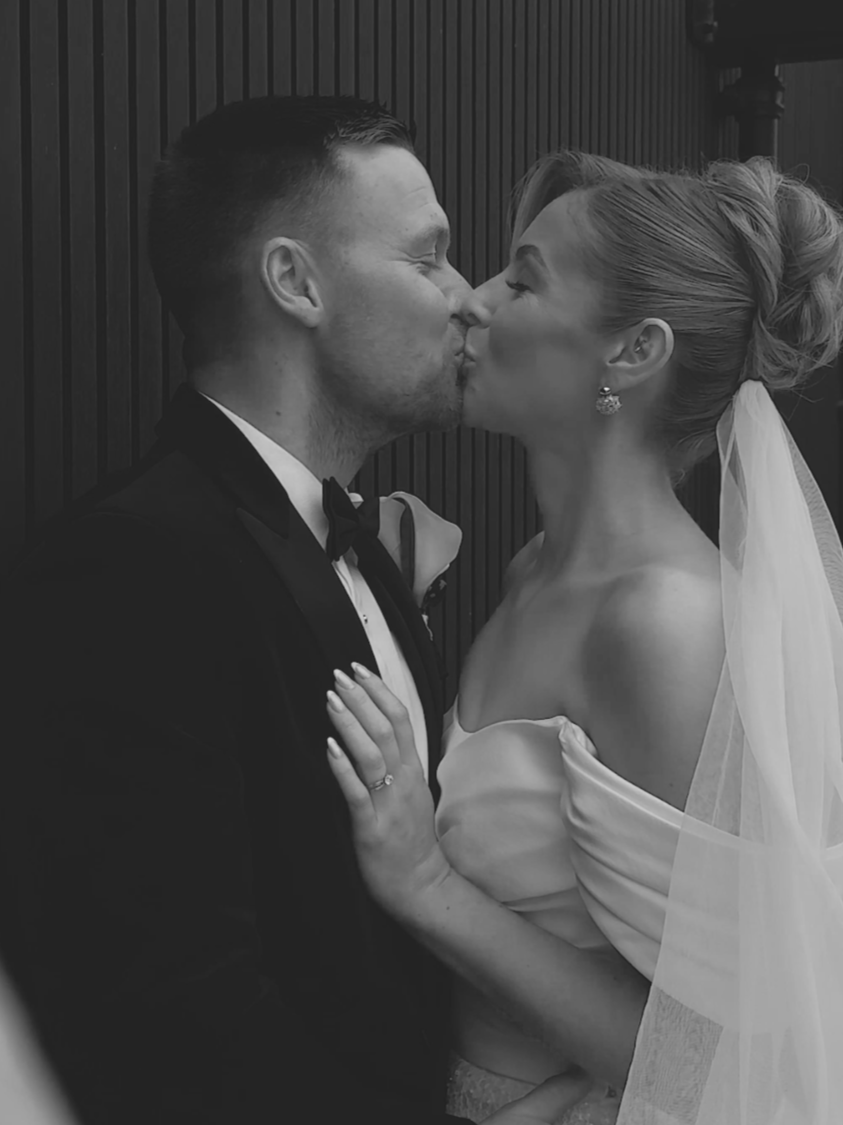 A black-and-white photograph of a bride and groom sharing a kiss, with the bride in a strapless wedding gown and veil, and the groom in a tuxedo with a boutonnière, standing close against a wooden wall.