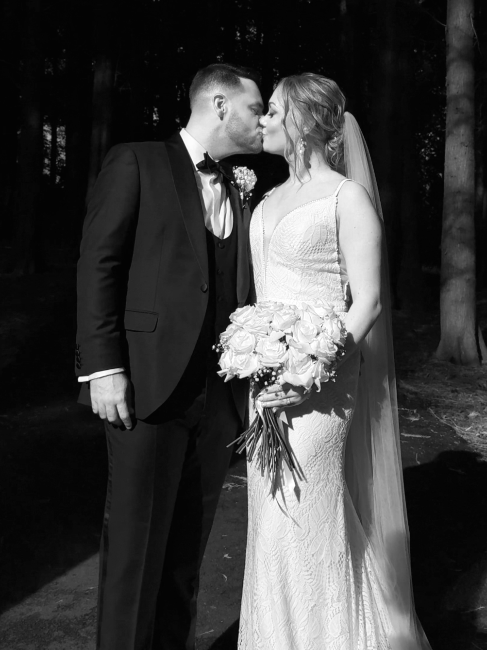 Black and white photo of a bride and groom kissing outdoors at night, with trees in the background. The groom is wearing a tuxedo with a bow tie, and the bride is in a lace wedding gown holding a bouquet of roses with a veil.