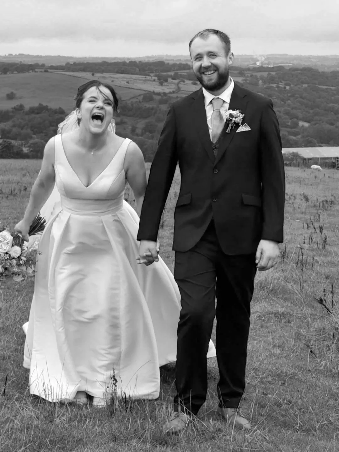 A happy bride and groom holding hands and walking in a field, with a scenic countryside in the background.