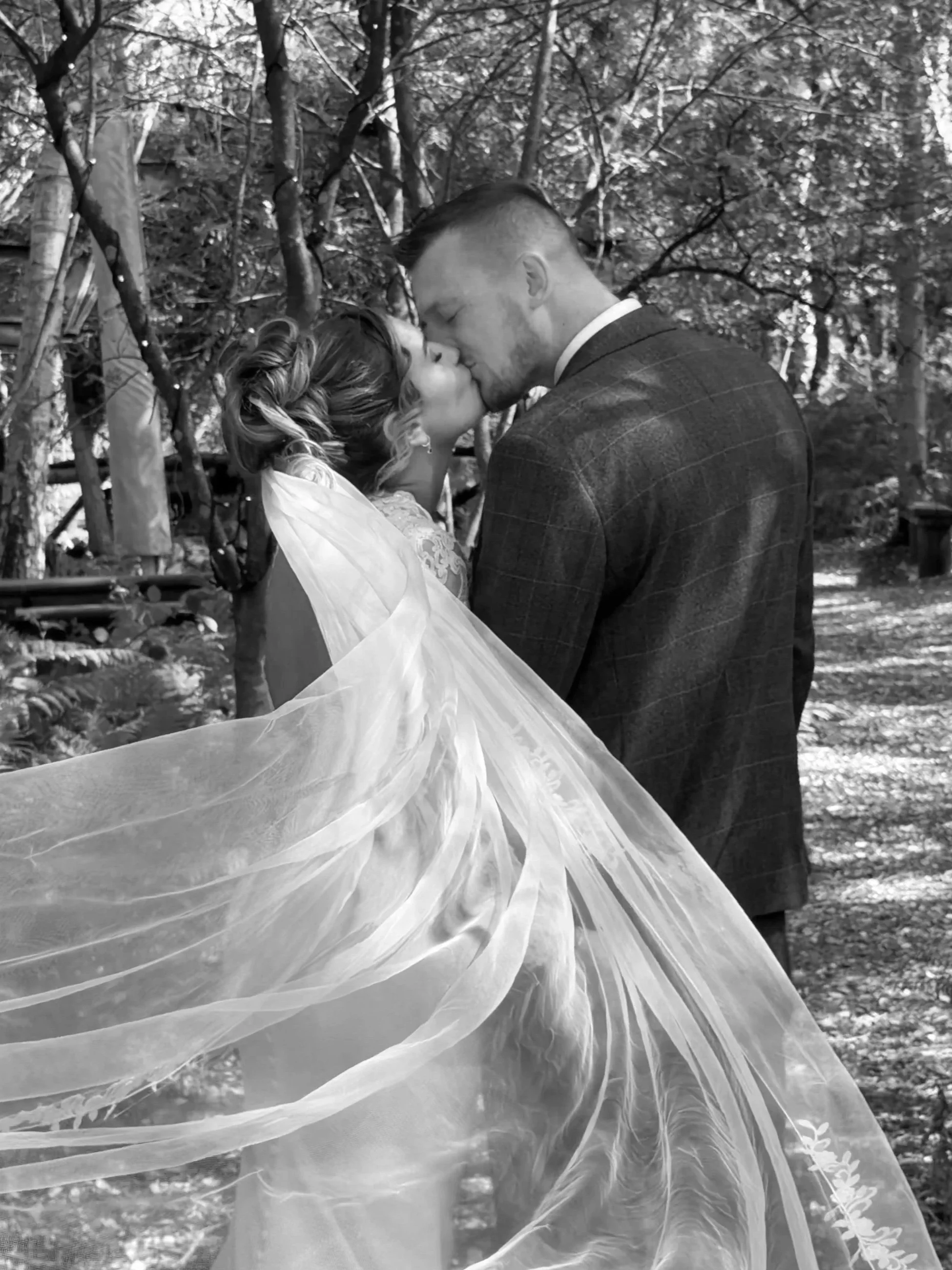 A bride and groom sharing a kiss outdoors, with the bride wearing a veil and the groom in a suit, surrounded by trees and natural light.