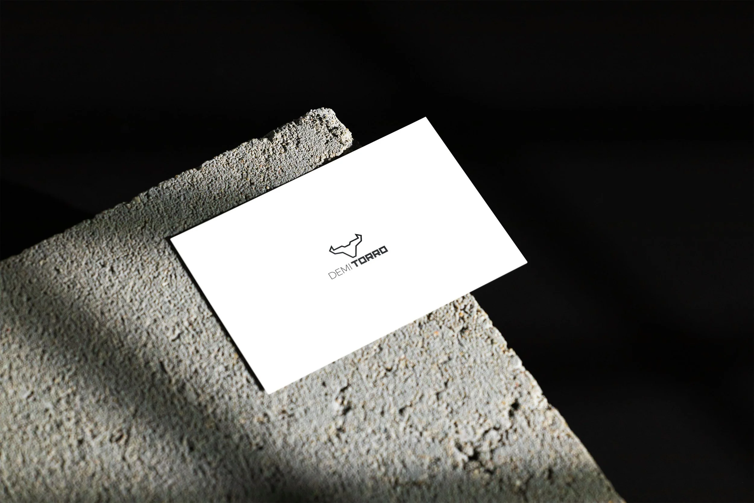 Business card with a minimalist logo and text 'DEMITORRO' resting on a textured concrete block against a dark background.