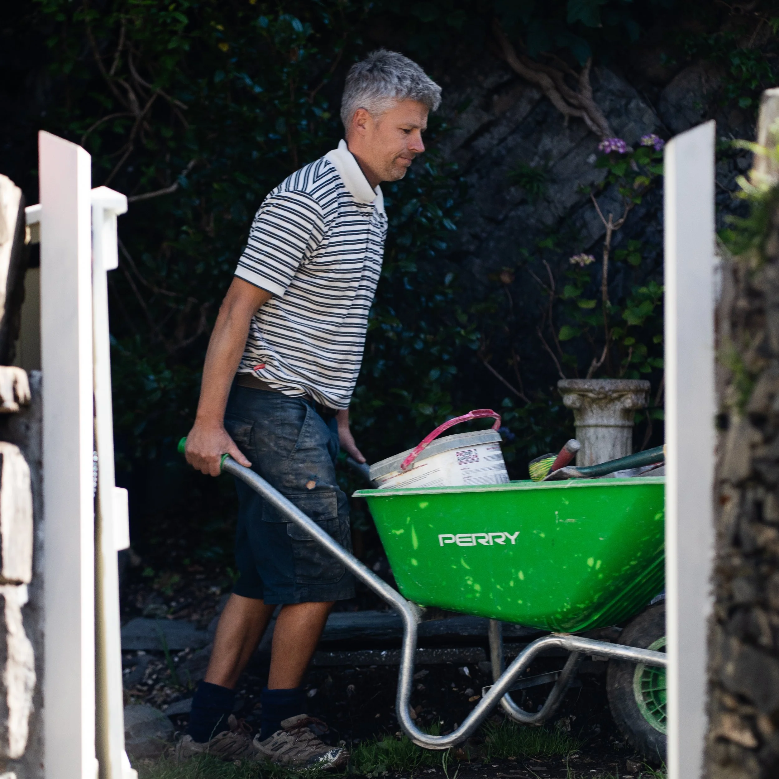 Man with gray hair pushing a green wheelbarrow with gardening tools and supplies in a garden.