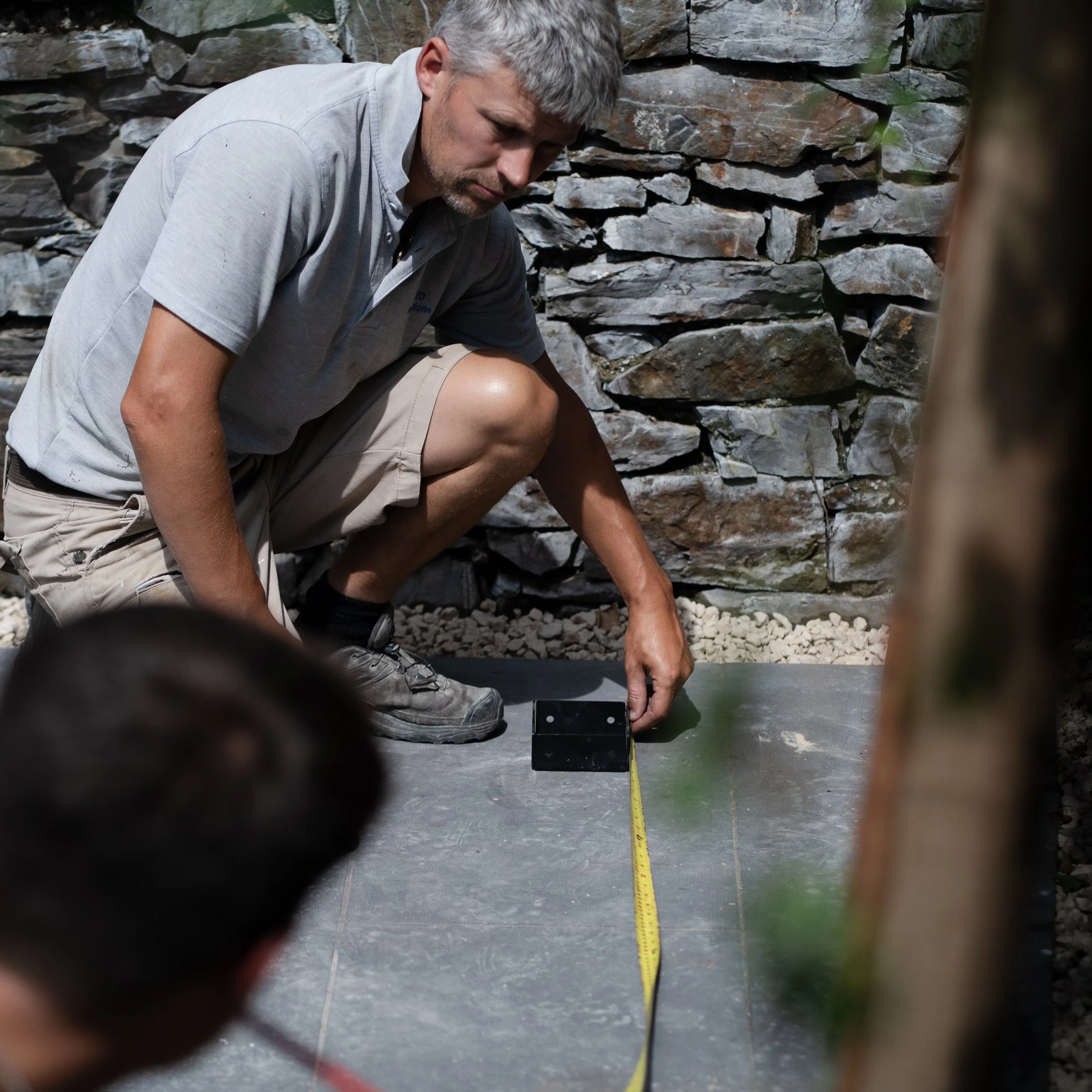 A man in a gray shirt and beige shorts kneels on a concrete surface, measuring with a tape measure beside a stone wall.