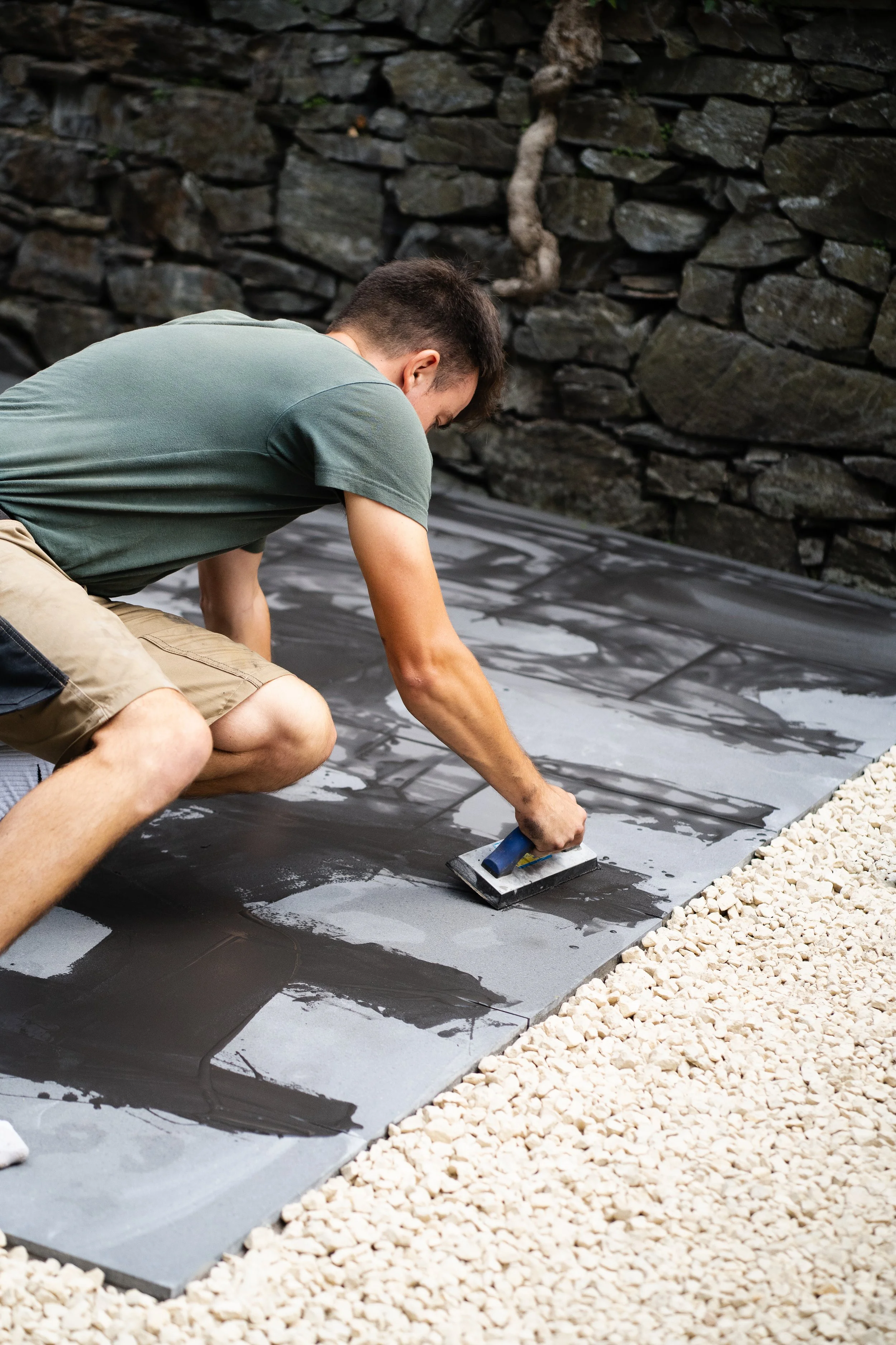 A man wearing a green t-shirt and beige shorts cementing large black tiles on the ground, with white gravel to the right and a stone wall behind.