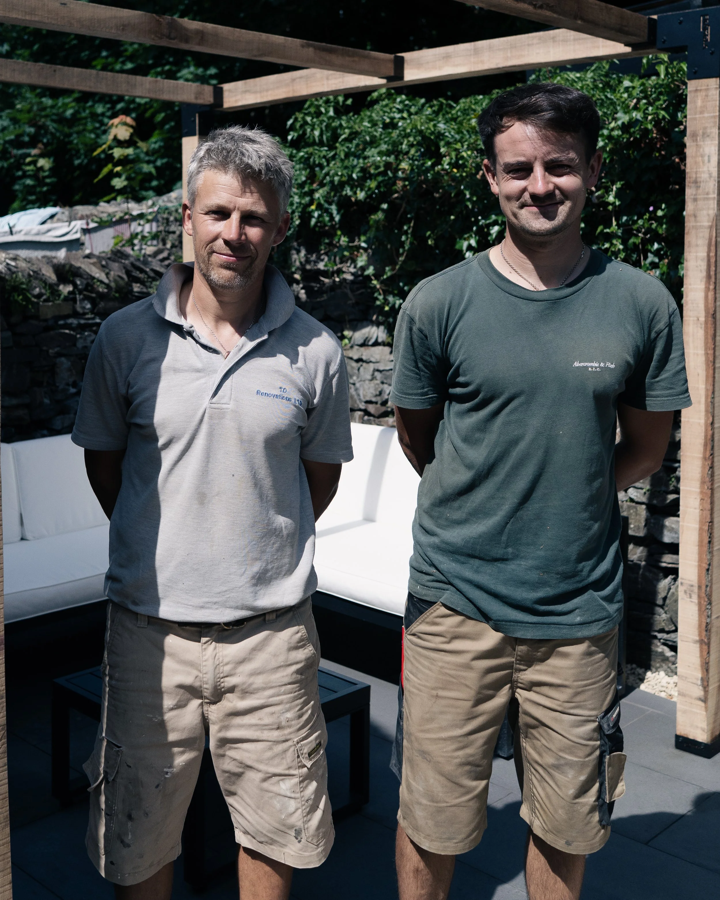 Two men standing outdoors with a wooden structure and greenery in the background, both smiling and wearing casual t-shirts and khaki shorts.