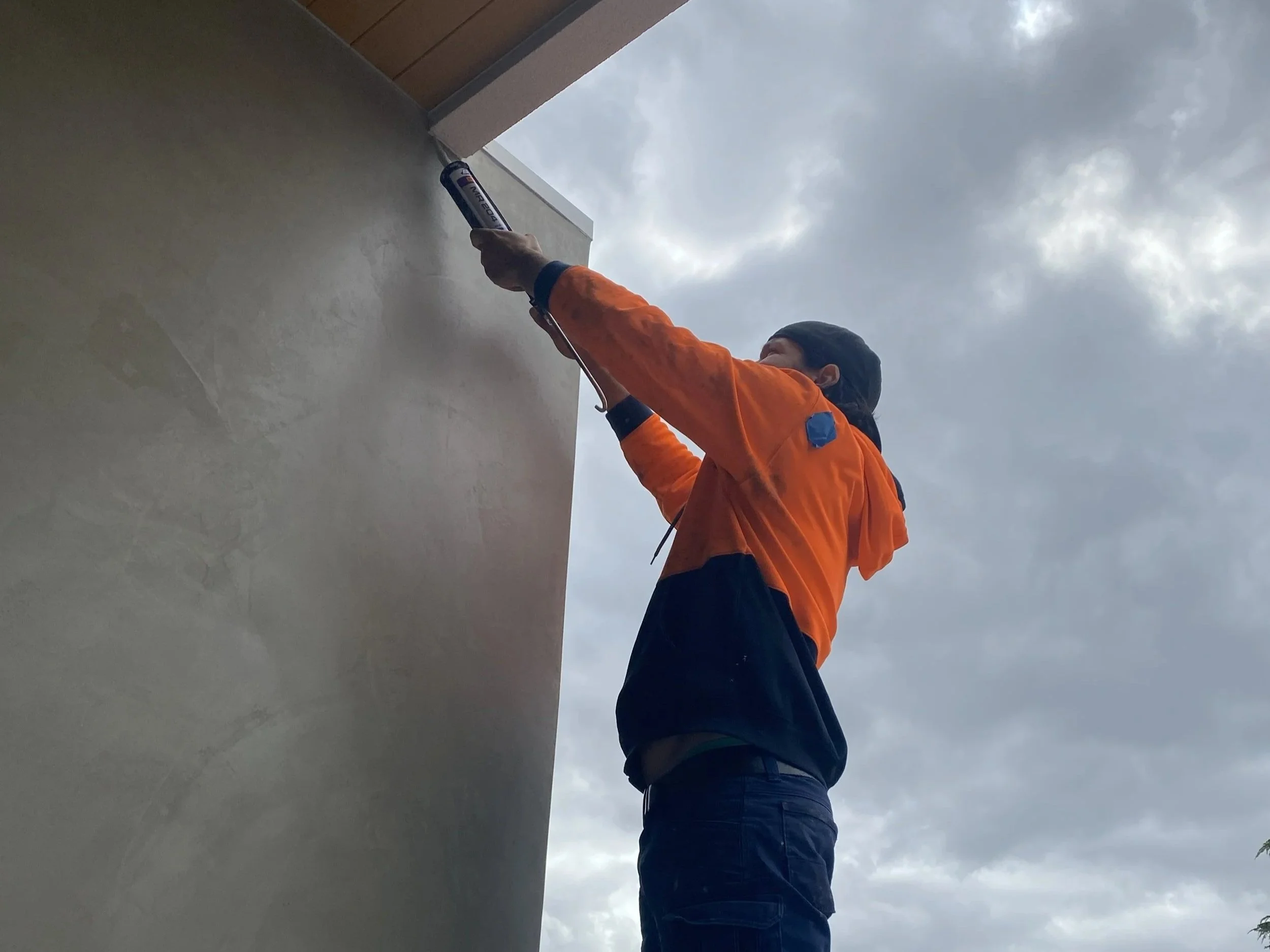 Person in orange jacket applying a sealant or adhesive with a caulking gun to the edge of a building or wall under a cloudy sky.