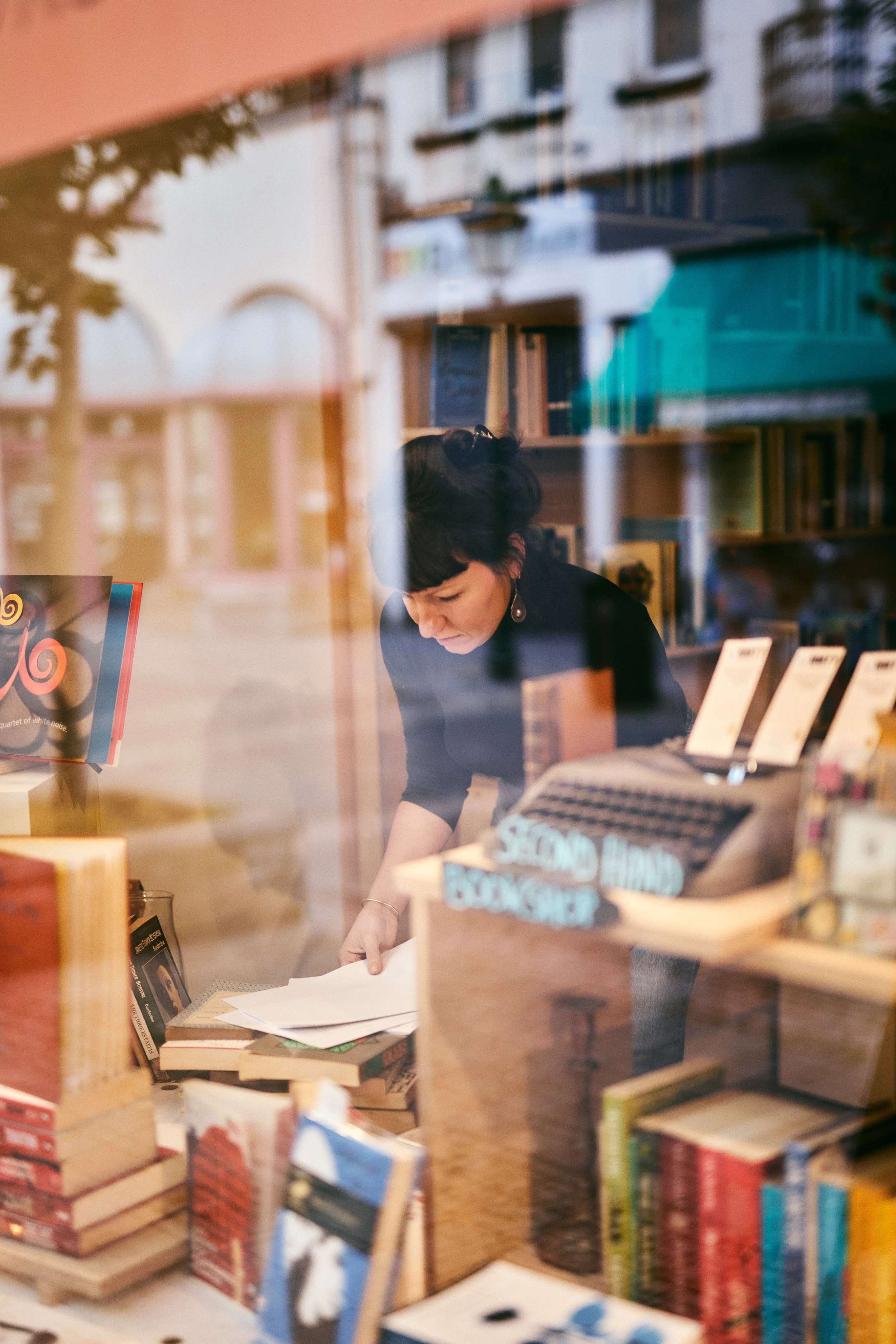 A woman with short dark hair and earrings is looking down while working with papers inside a bookstore or library, seen through a reflective window with books on display.