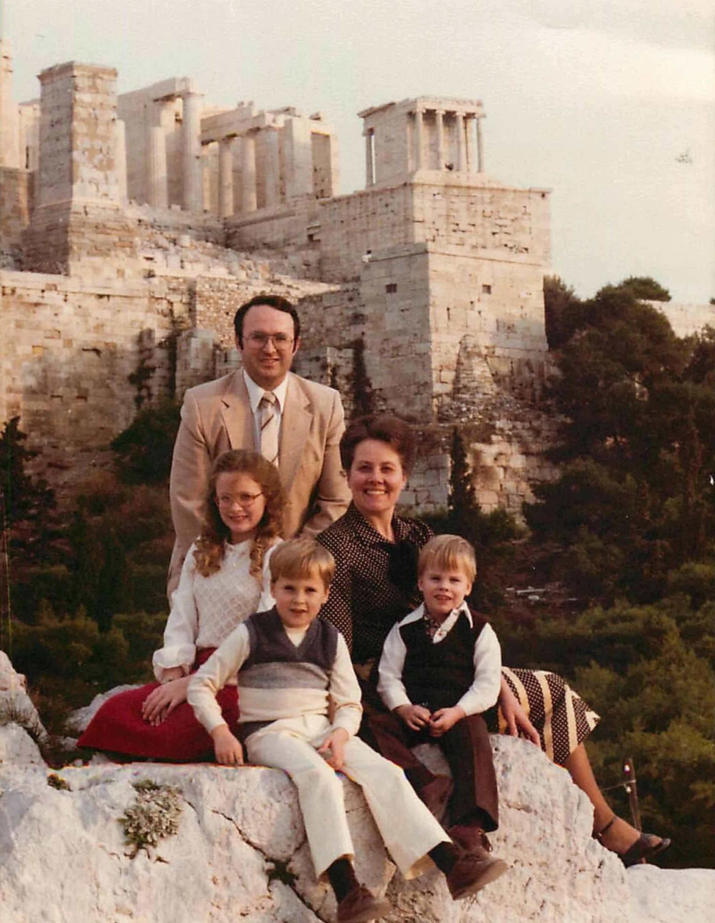 A family of six posing on a rocky ledge in front of the Parthenon in Athens, Greece, during daytime. The family includes two adults and four children, with the ancient Greek temple in the background.