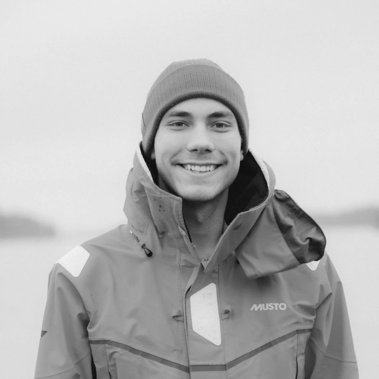 Black and white photo of a smiling man wearing a beanie hat and casual clothing, standing outdoors near water with a blurred background.