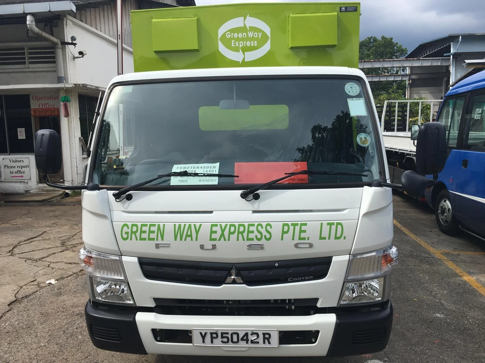 Front view of a white Mitsubishi Canter truck with the words 'GREEN WAY EXPRESS PTE. LTD.' written in green on the front. The truck is parked in a parking lot next to other trucks, with a green square container on top that has a white logo 'Green Way Express' at the top. There is a building to the left and another truck to the right.