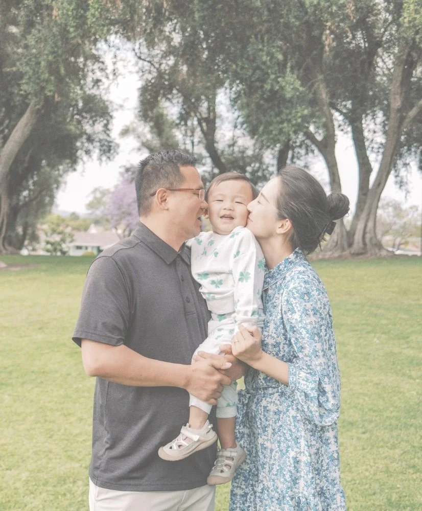 A family of three, including a father, mother, and young child, standing outdoors on a grassy field with trees in the background, enjoying a happy moment together.