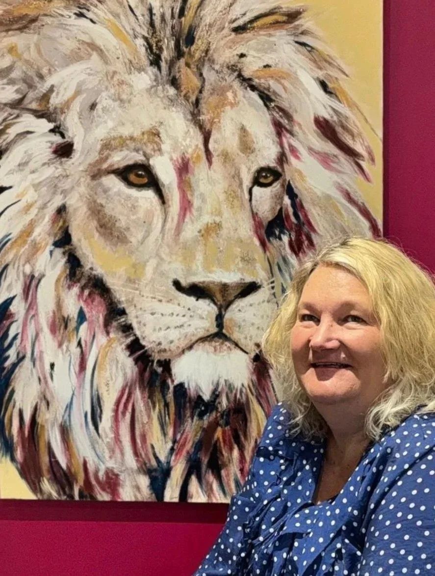 A woman with blonde hair smiling and wearing a blue polka dot shirt posing in front of a large colorful painting of a lion.