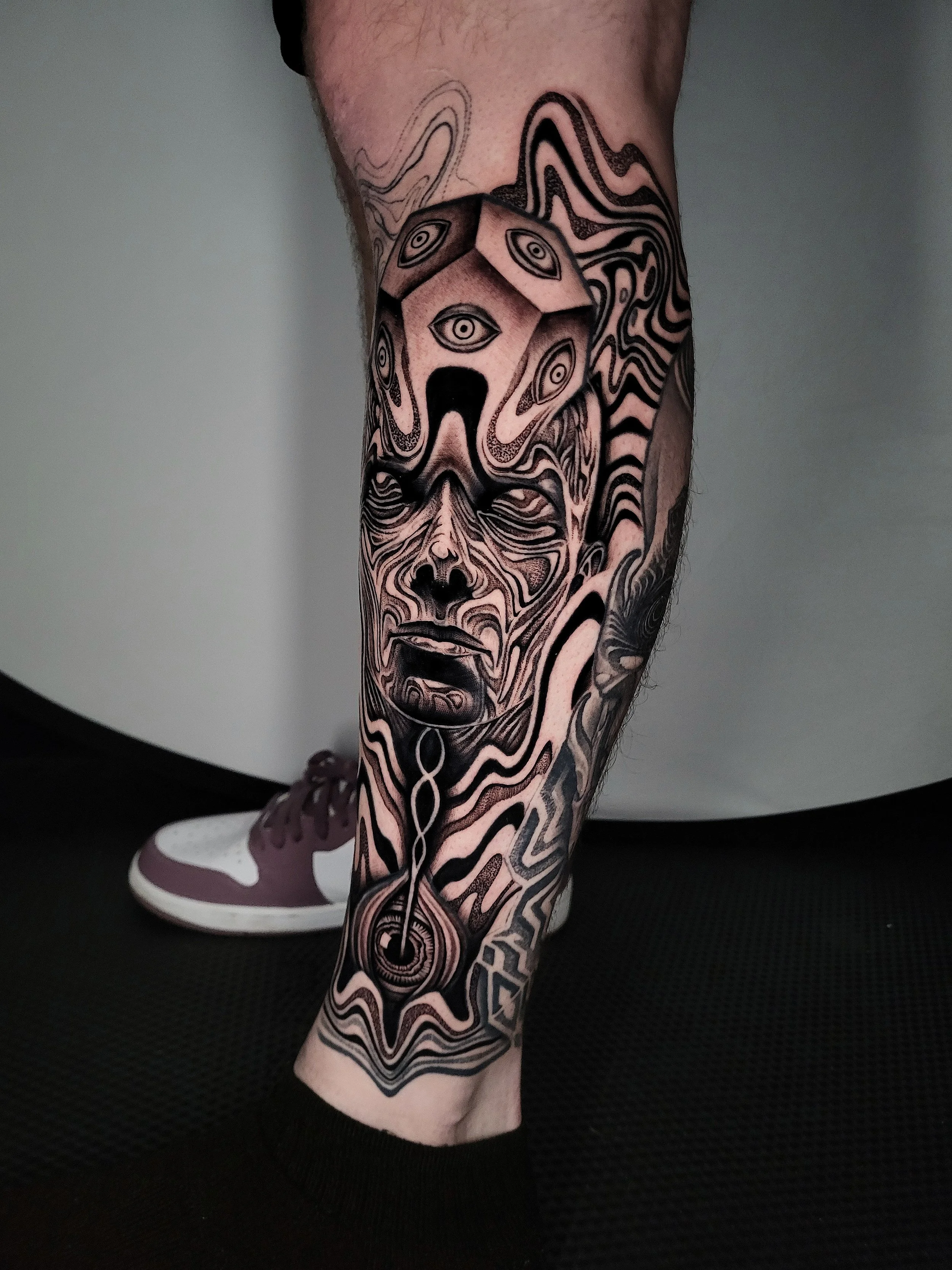 Black and gray tattoo of a surreal, abstract face with multiple eyes, flowing lines, and intricate patterns covering a person's lower leg.