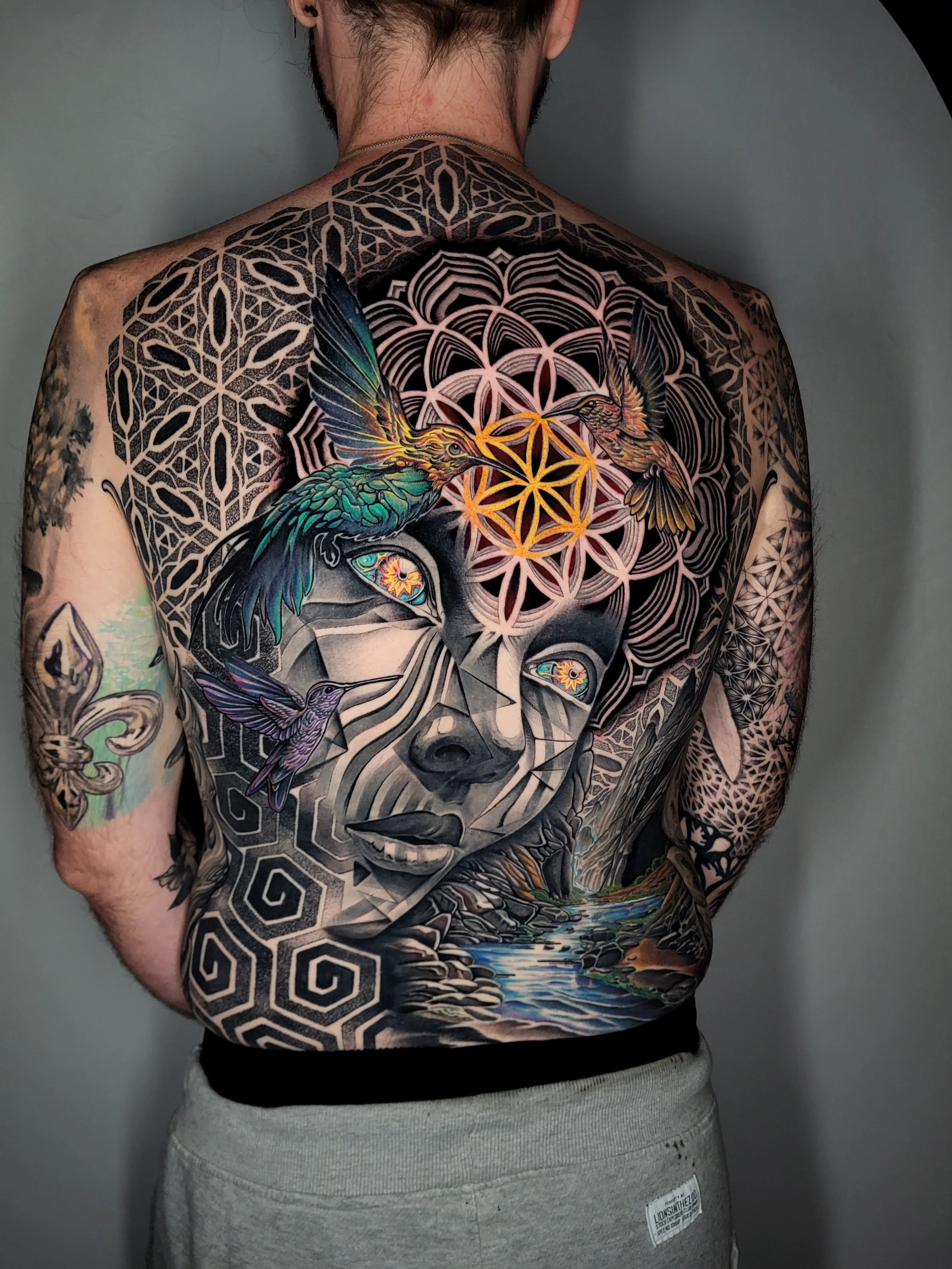 Colorful tattoo of a woman's face with geometric patterns, hummingbirds, and a landscape on a person's back.