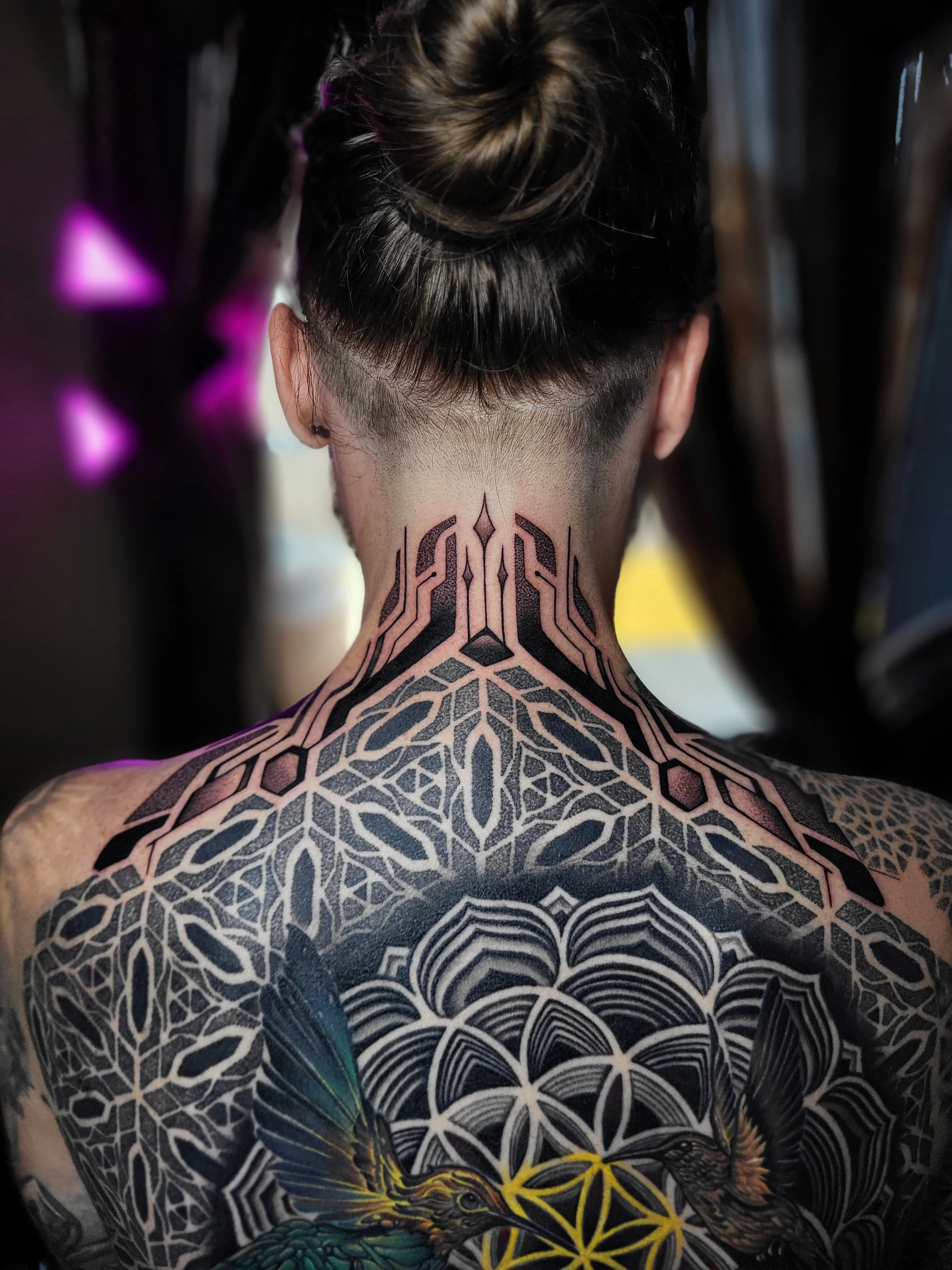 Back view of a person with tattoos on their neck and shoulders, including geometric patterns, birds, and a flower design.