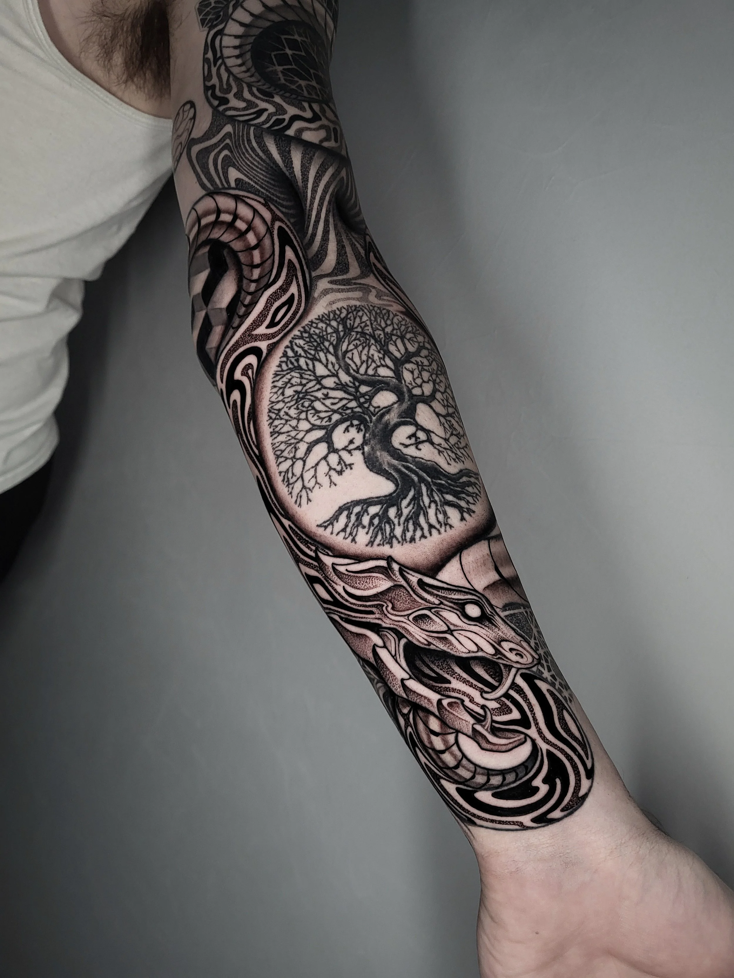 Black and gray tattoo sleeve featuring a large tree with expansive branches and roots, intricate patterns and designs surrounding the tree extending down the arm.