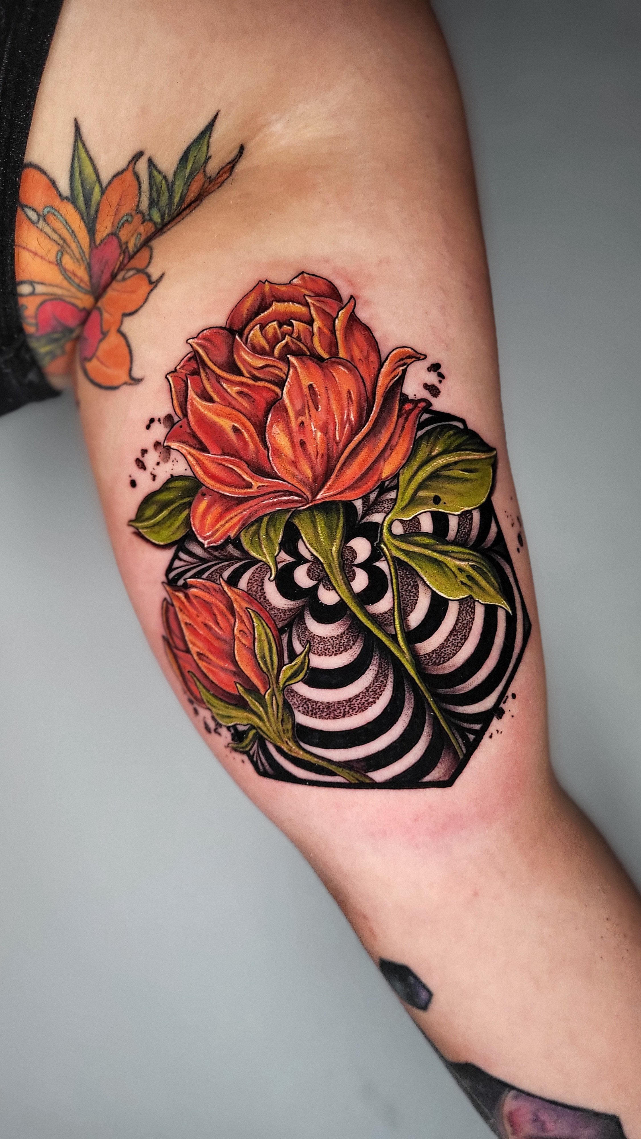 Colorful tattoo of orange roses with green leaves over black and white geometric and spiral background on person's arm.