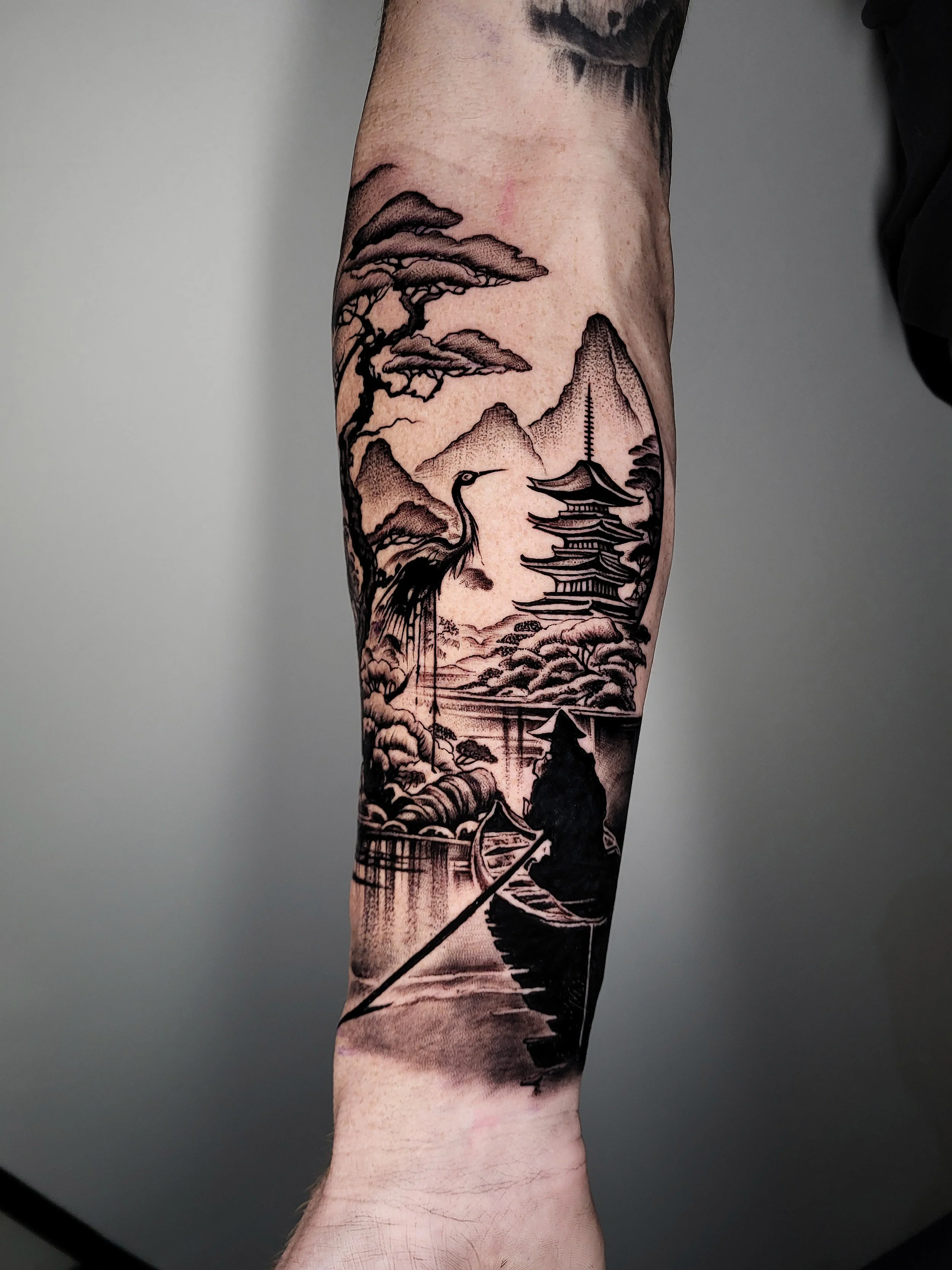 Black and gray tattoo of a Japanese landscape scene with a pagoda, mountains, pine trees, a heron, a person rowing a boat, and a bridge.
