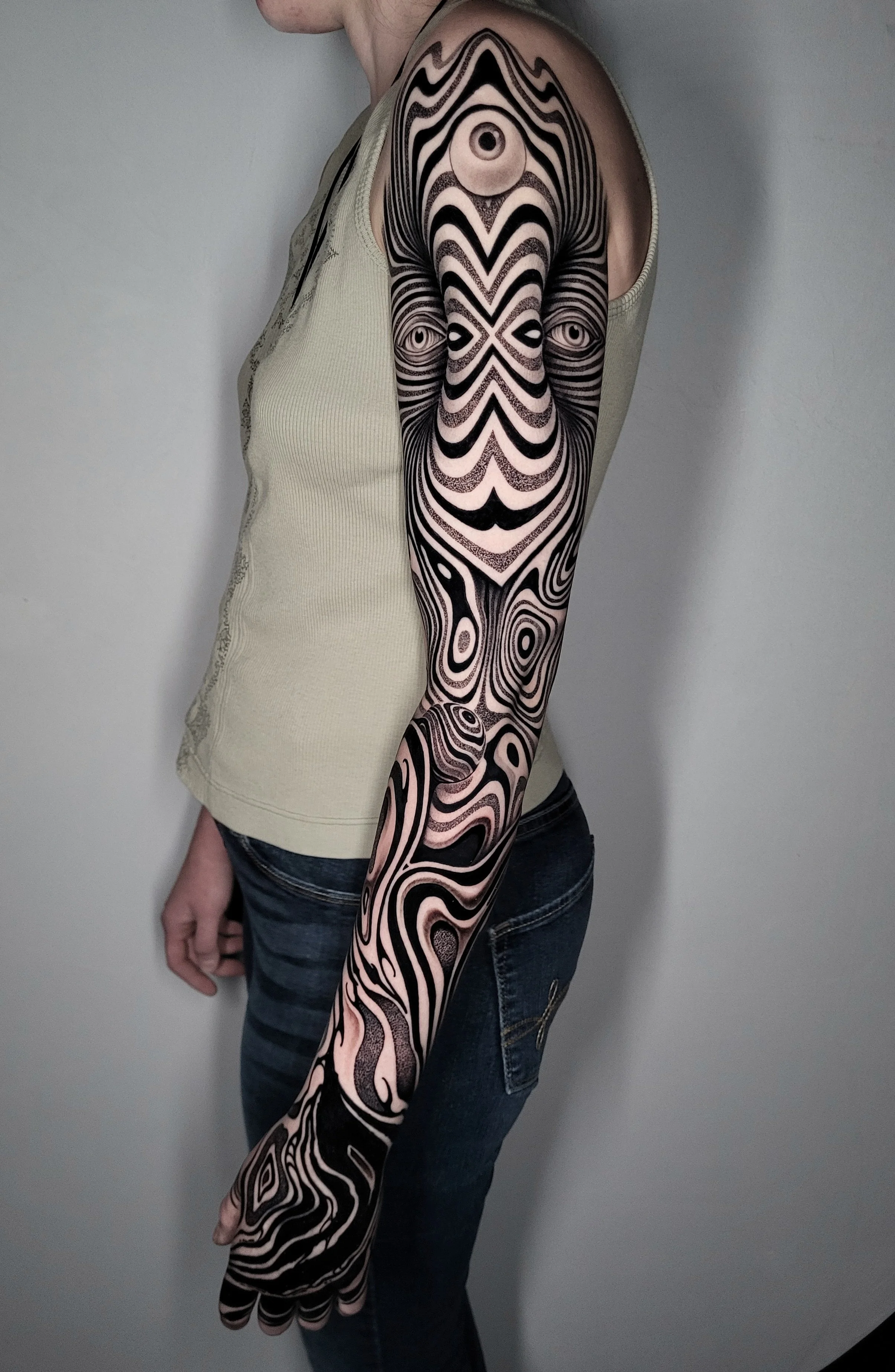 A person with a black and beige abstract patterned tattoo on their entire arm, standing sideways against a plain wall.