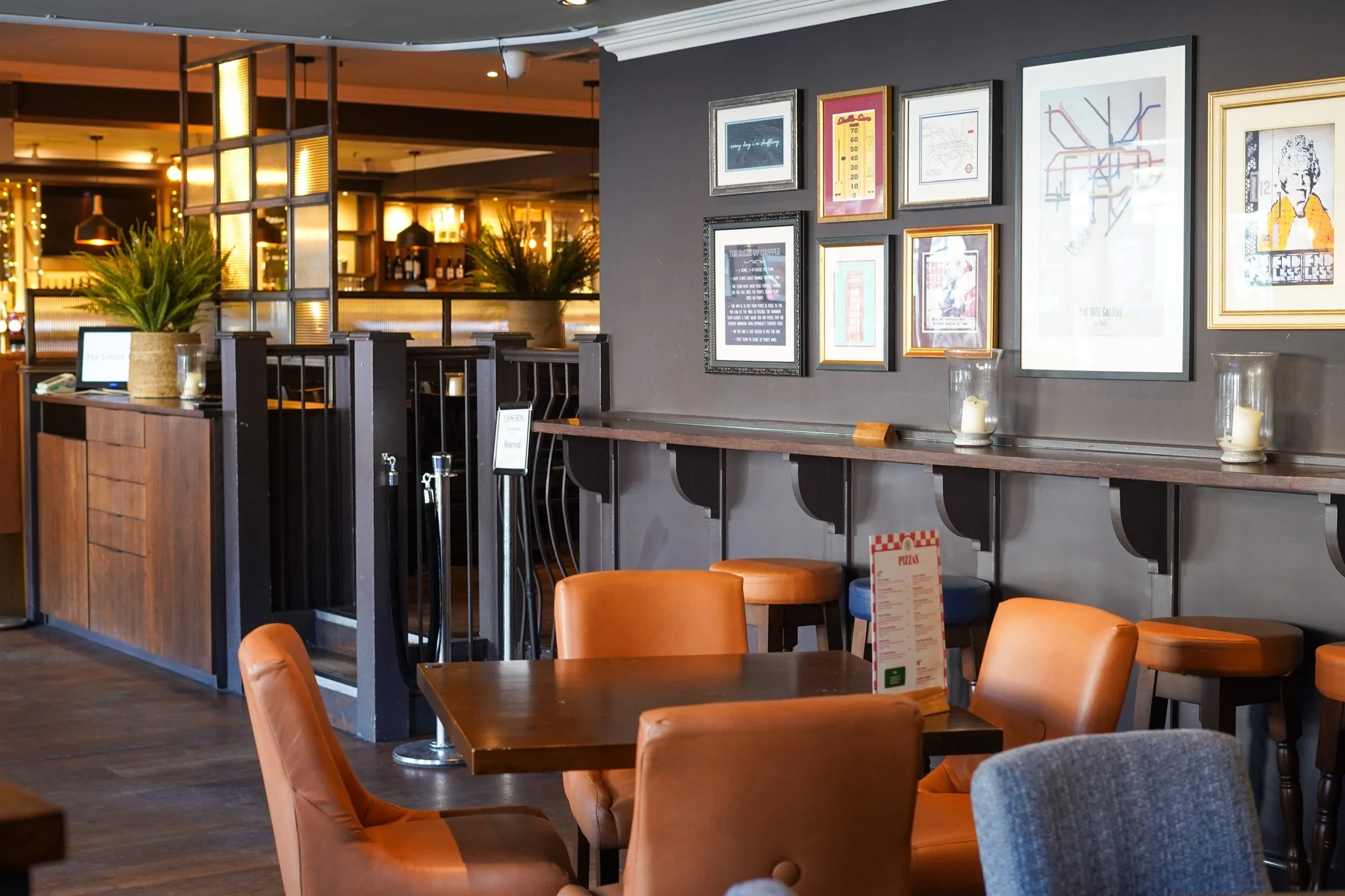 Interior of a cozy restaurant or cafe with tan and gray chairs, wooden tables, and framed artwork on dark gray walls, illuminated by warm lighting.