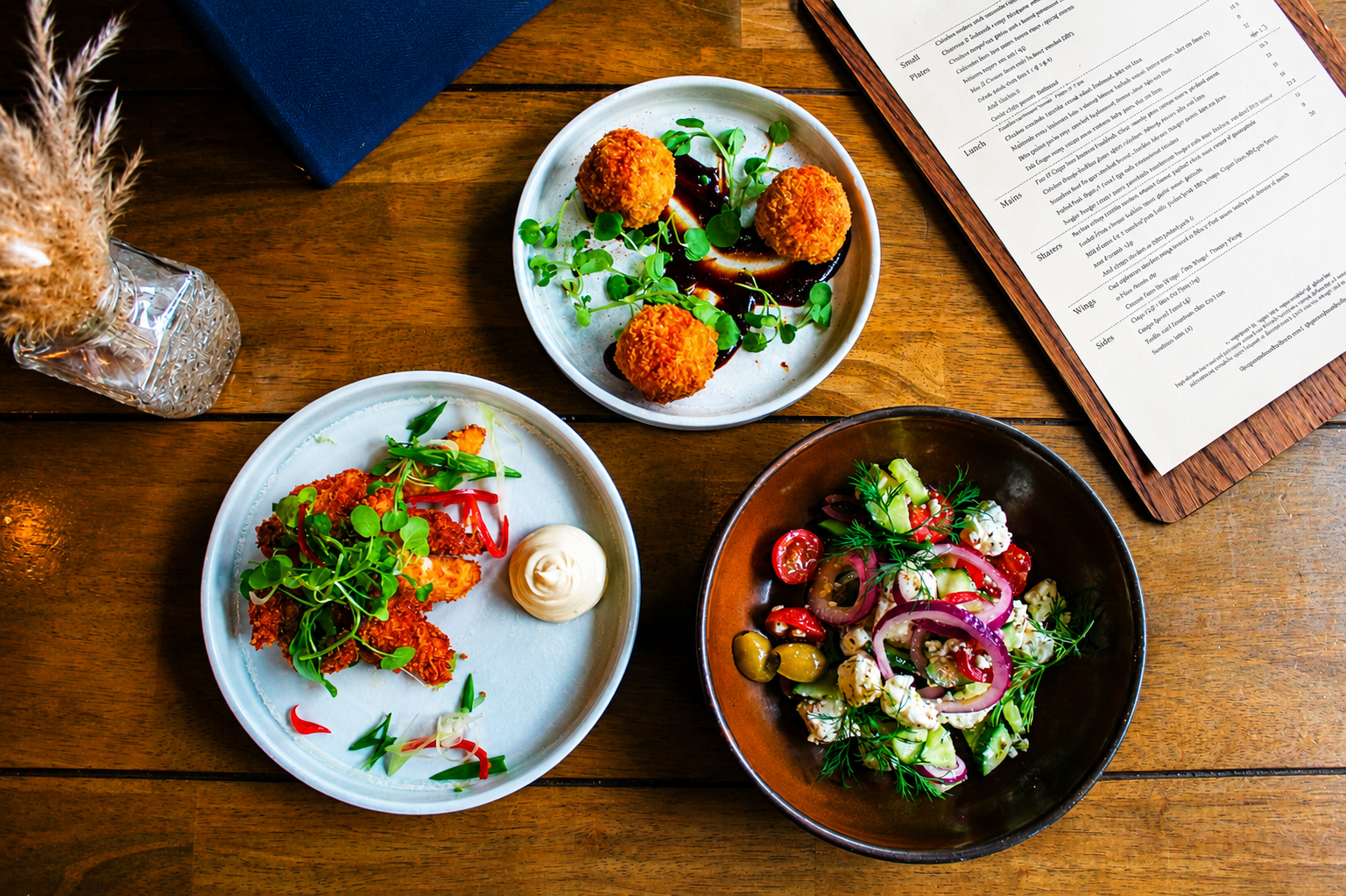 Three plates of food on a wooden table, with a menu and a vase of dried grass. The plates feature a salad with tomatoes and herbs, fried ball appetizers with sauce and greenery, and a dish with fried chicken, greens, and sauce.