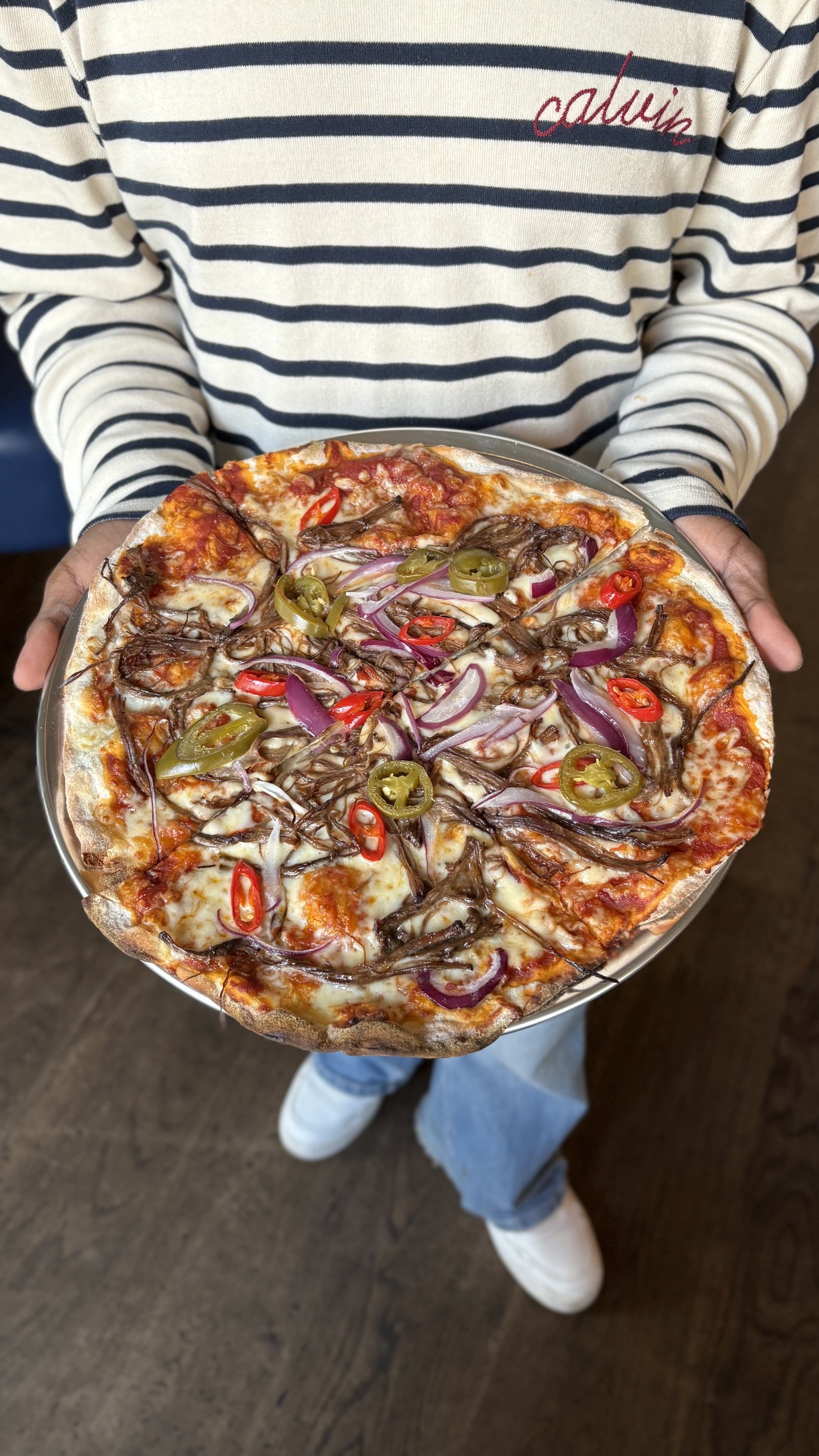 Person in a striped shirt holding a pizza with red and green peppers, onions, and shredded cheese.